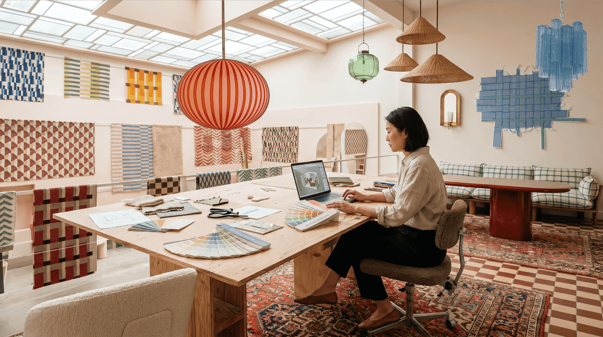 Interior designer working in SketchUp on a laptop, with Benjamin Moore and Sherwin-Williams paint fan decks and color chips fanned across a plywood worktable in a bright, fabric-filled studio.