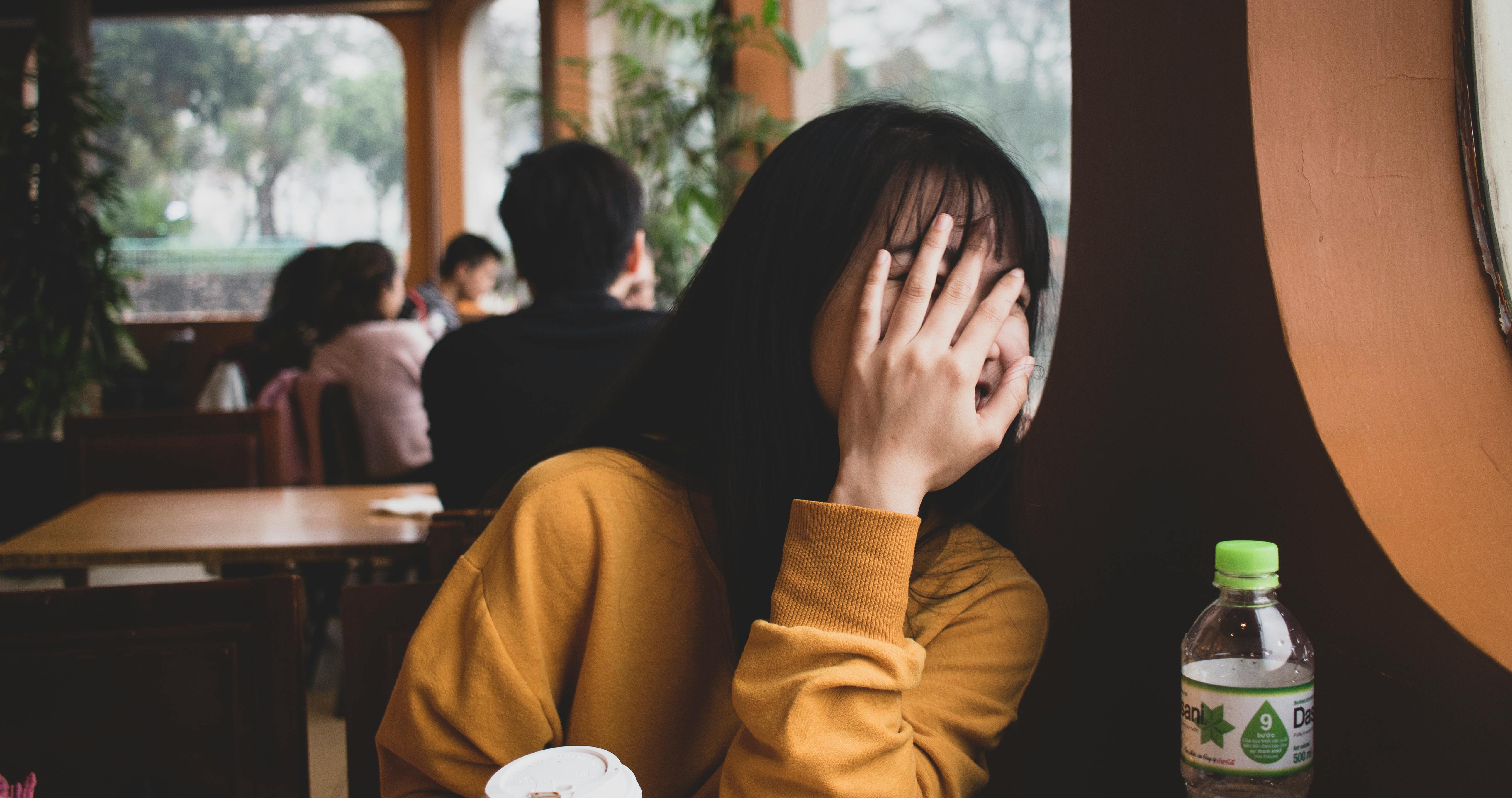 A woman is shying away from the camera, hiding her face while nervously laughing, while seating in a public space at a cafe