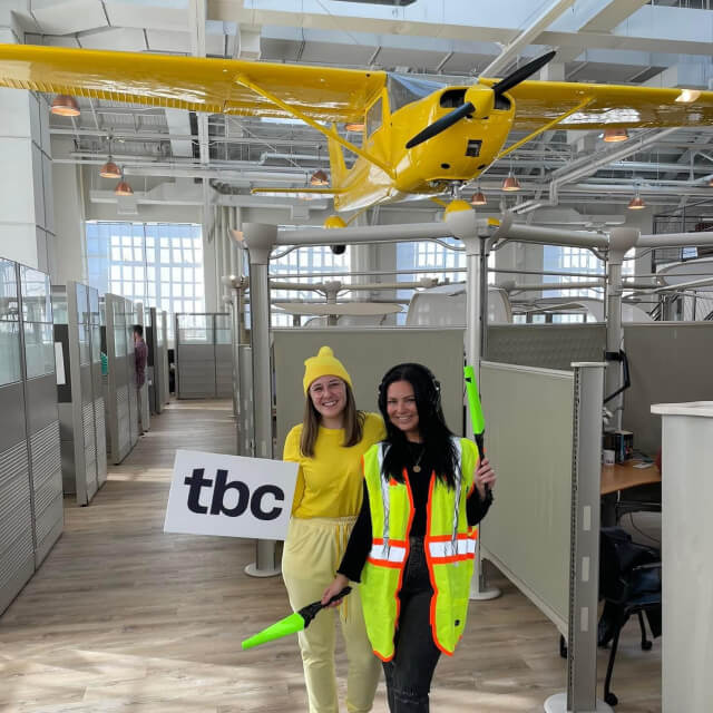 Two people pose indoors under a yellow plane model; one wears yellow and holds a "tbc" sign, the other wears a safety vest and holds green batons.