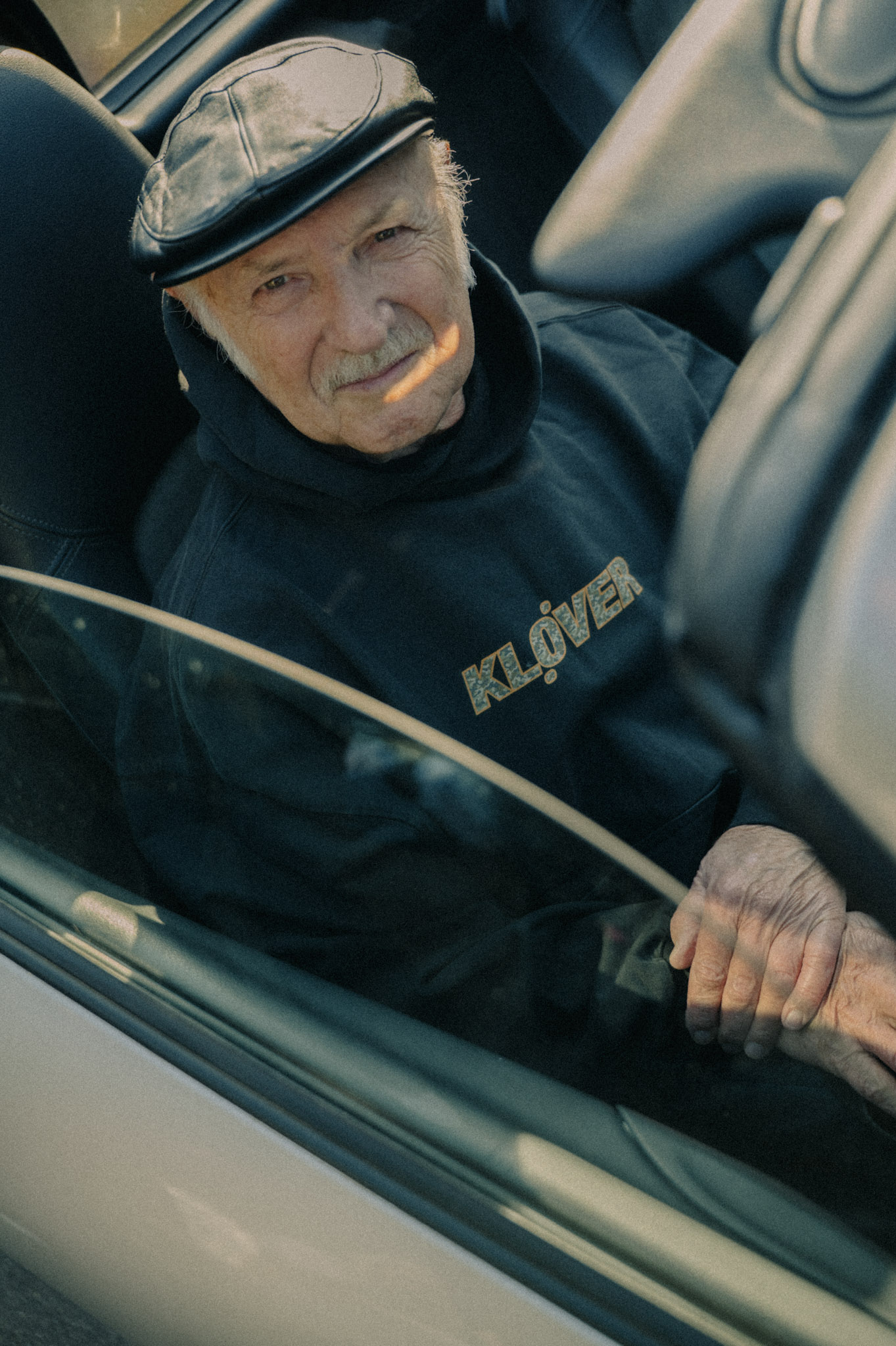 Old man dressed in Kløver clothing, sitting in a convertable car. 