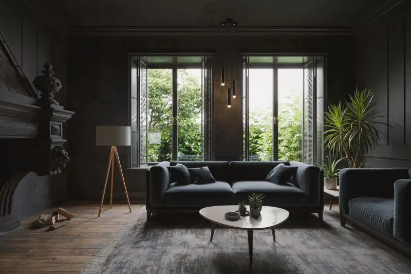 A high-quality 3D render of a dark, luxurious living room with a deep blue velvet sofa, tripod lamp, and bright windows looking out onto foliage