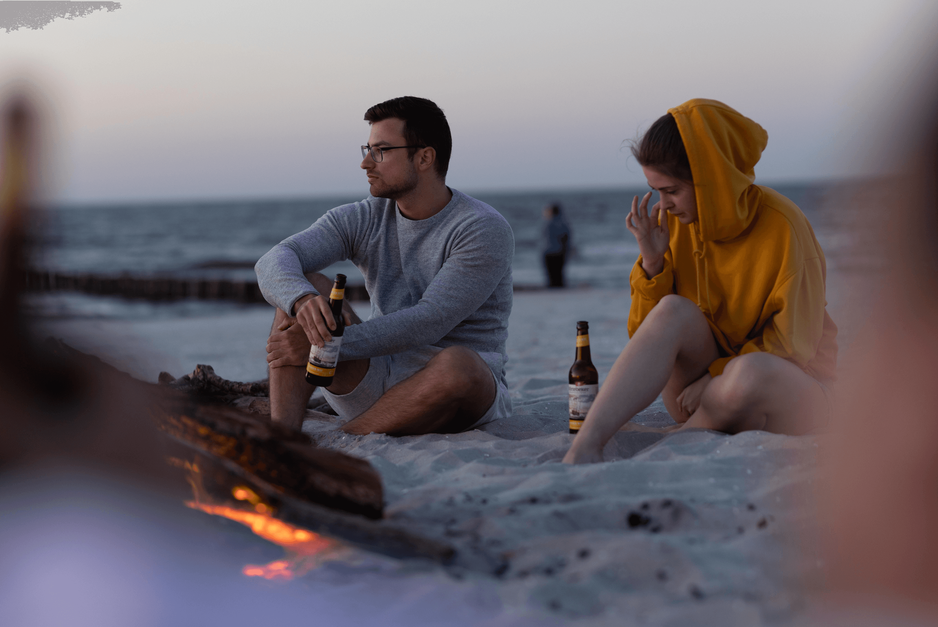 Couple with beer bottles on beach at dusk.