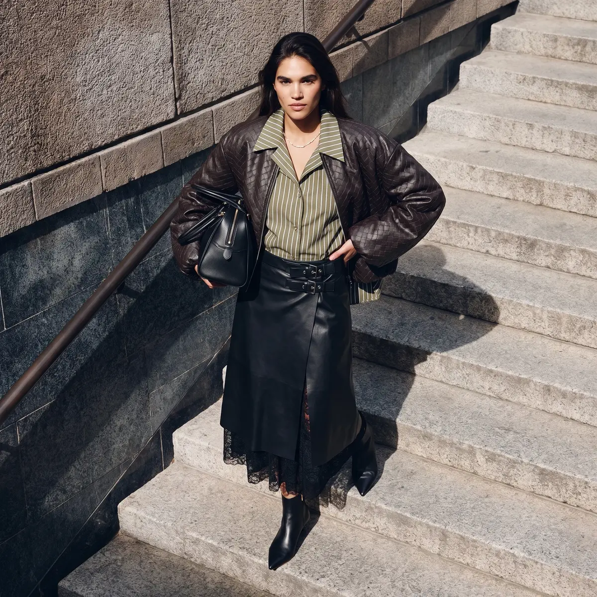 Modella sulle scale con giacca in pelle intrecciata, camicia a righe e gonna in pelle