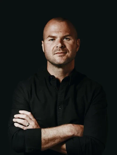 A confident individual with a shaved head, wearing a black button-up shirt, stands against a dark background, arms crossed, conveying a professional and self-assured presence.