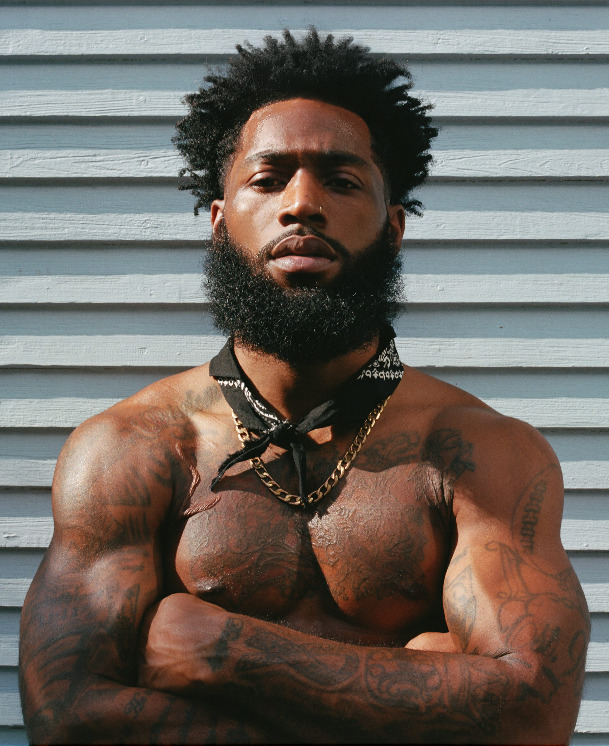 A heavily tattooed man stands shirtless with arms crossed against light blue wood siding, direct gaze toward camera.