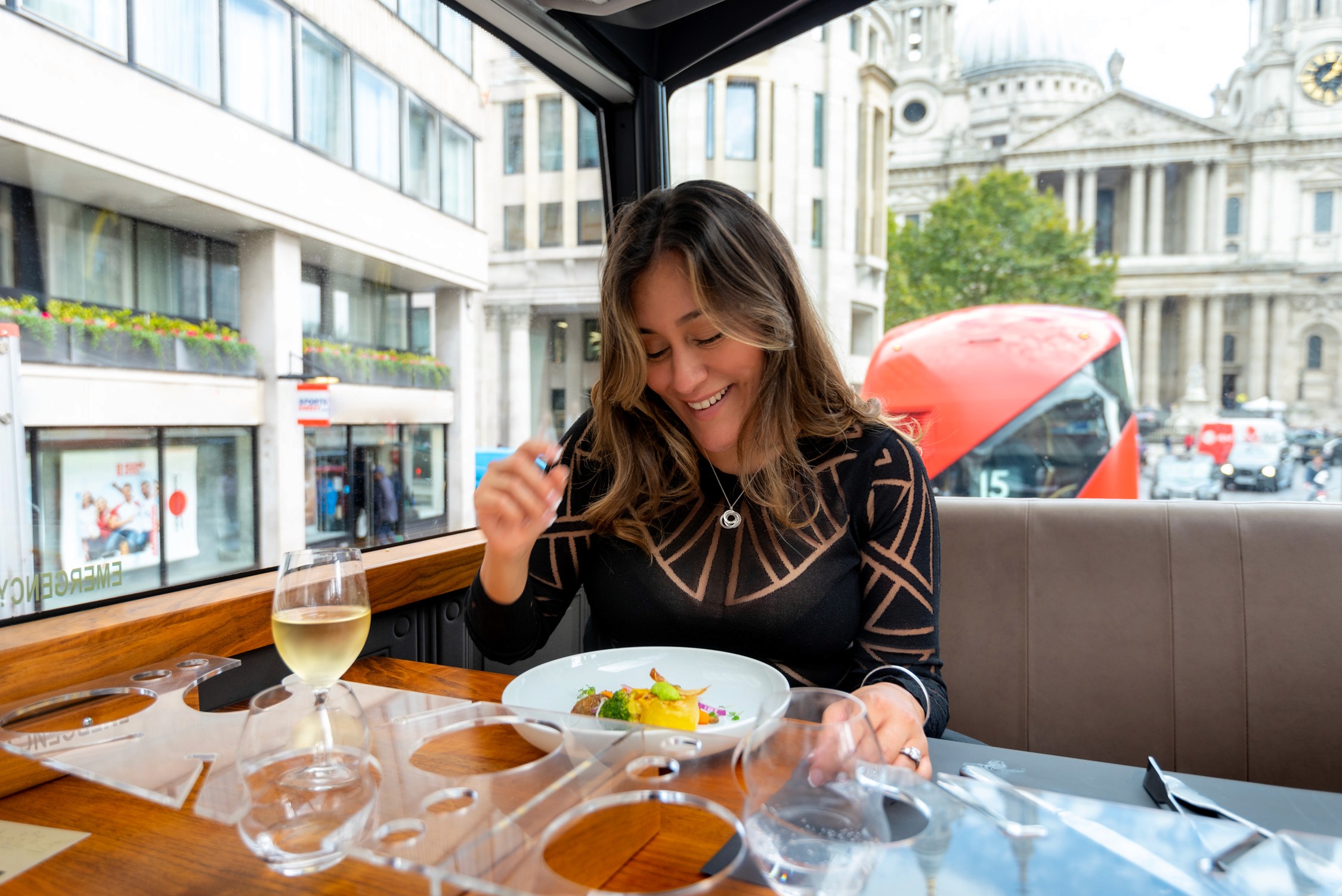 A woman eats a dish of pasta and salad at a table in an open-air bus tour vehicle overlooking a city street.