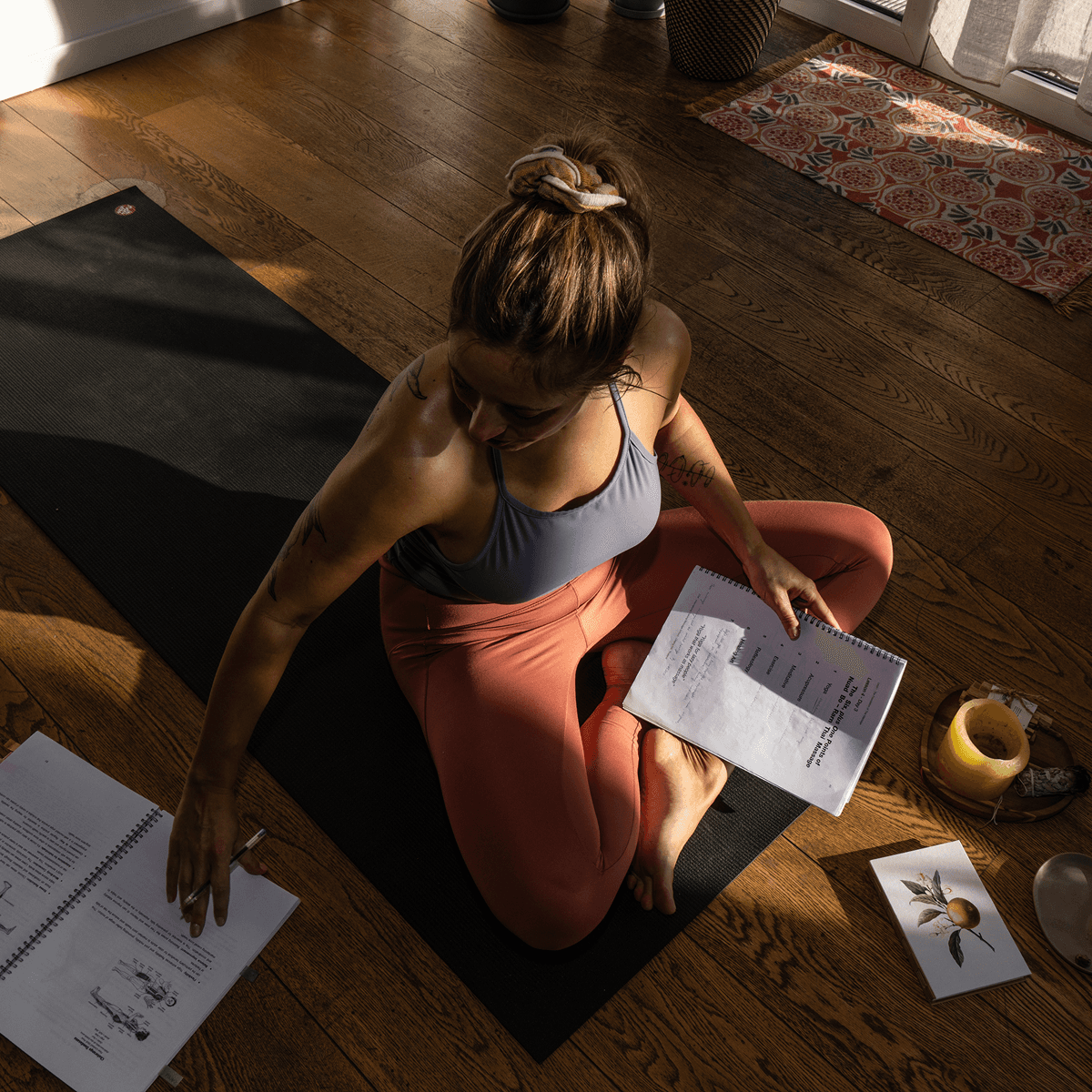 A woman sits on a yoga mat in a sunlit room, multitasking by reviewing documents spread around her, while in a relaxed yet focused posture.