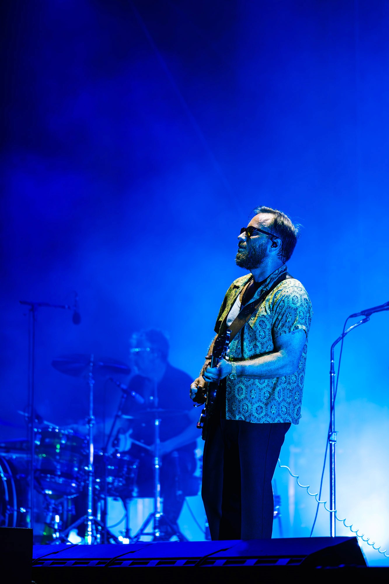 Guitariste en chemise à motifs sous une lumière bleue intense, batteur en arrière-plan flou