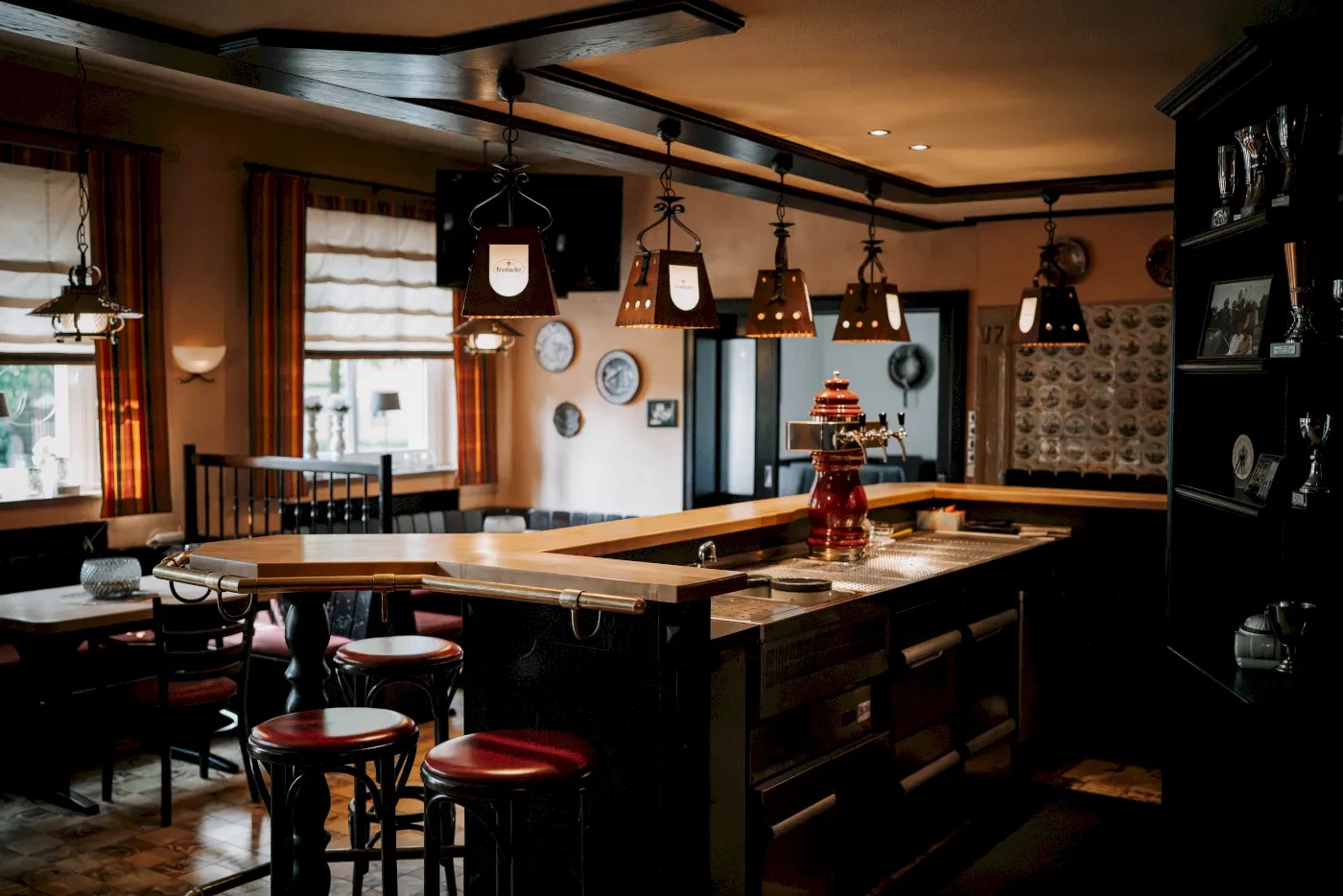 Gemütliche Kneipe mit Bar, Holztischen und Stühlen. Blick auf die Theke, Fenster, Kronleuchter und Dekoration.