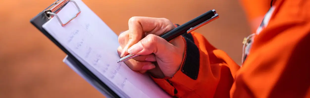 A person in an orange jacket is writing on a clipboard, possibly documenting information related to a risk engineering assessment or using a loss control app for an insurance inspection platform.