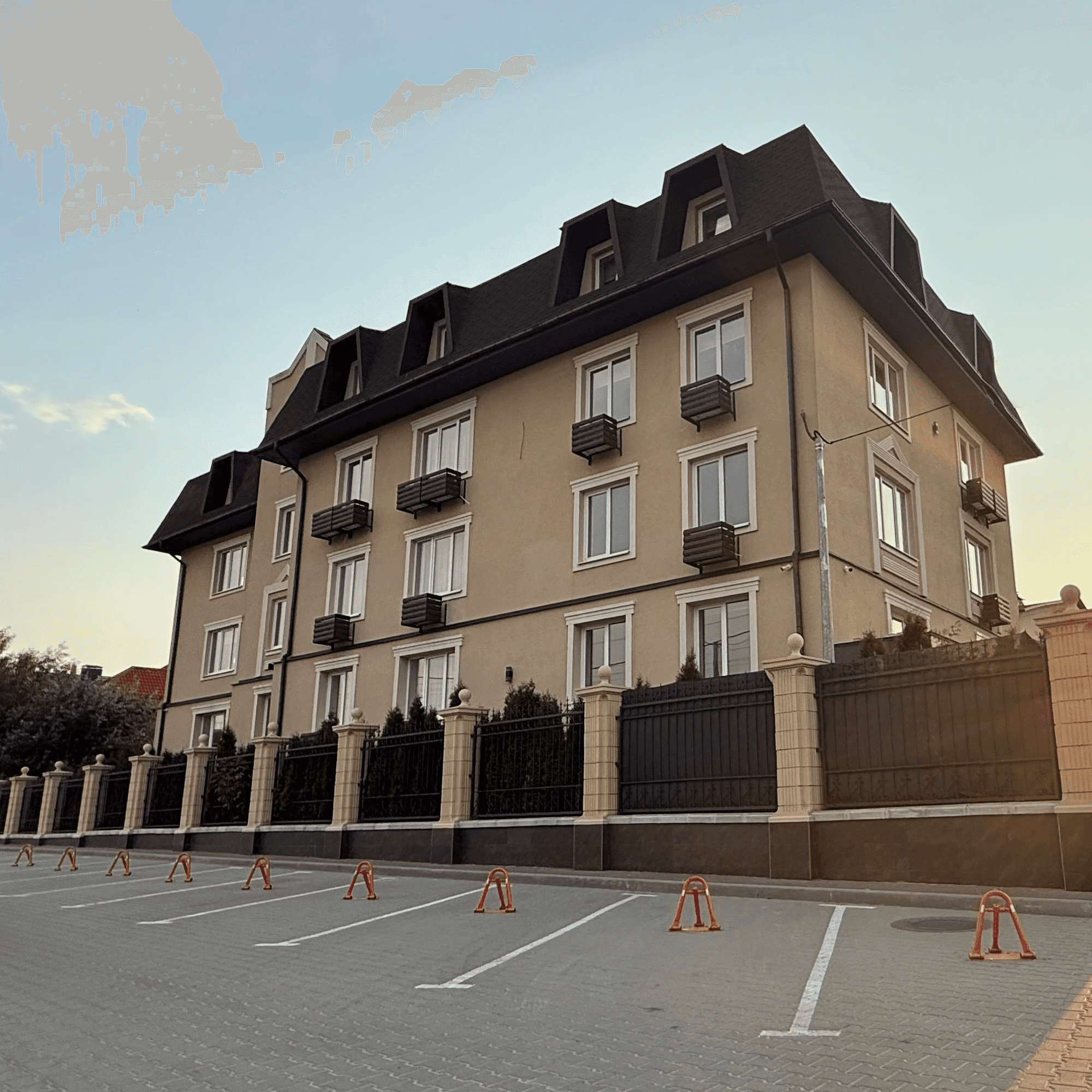 Фасад чотириповерхового будинку з декоративними балконами, високим дахом і огородженою територією