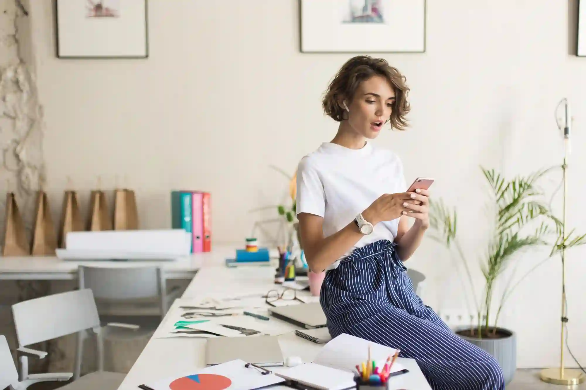 A woman leaning on a messy desk while excitedly checking her phone, likely tracking sales or campaign performance.