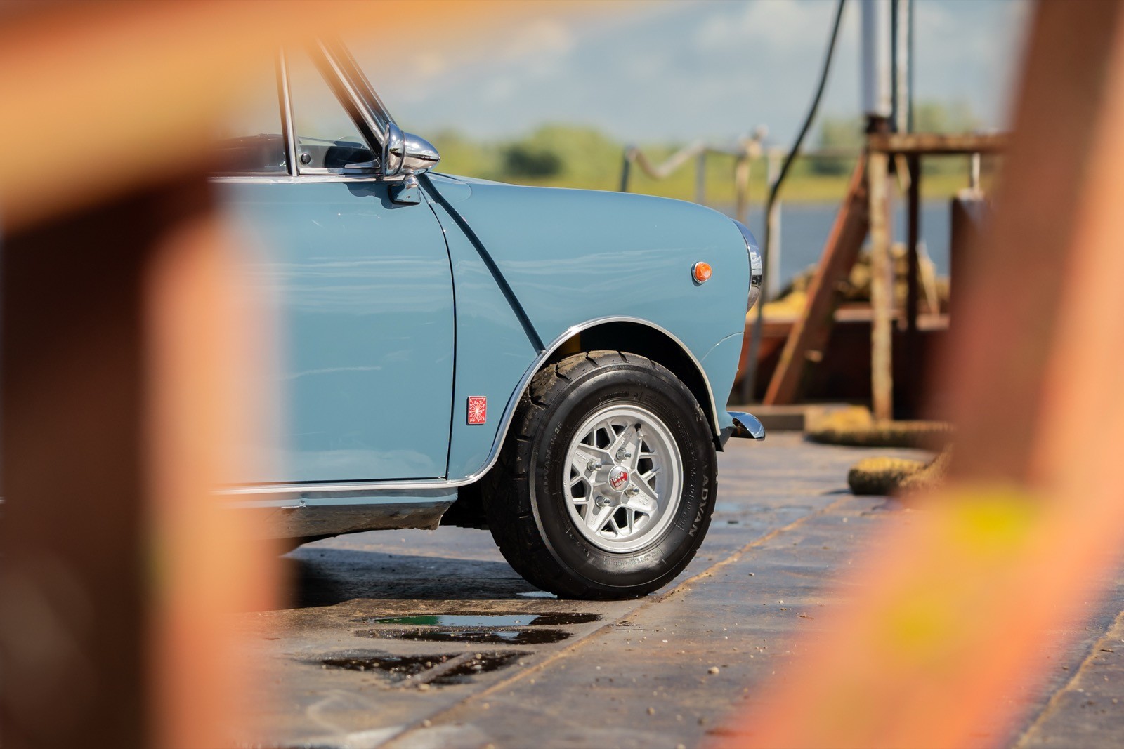 A close-up view of a light blue car parked by a wooden structure near water, framed by wooden beams.