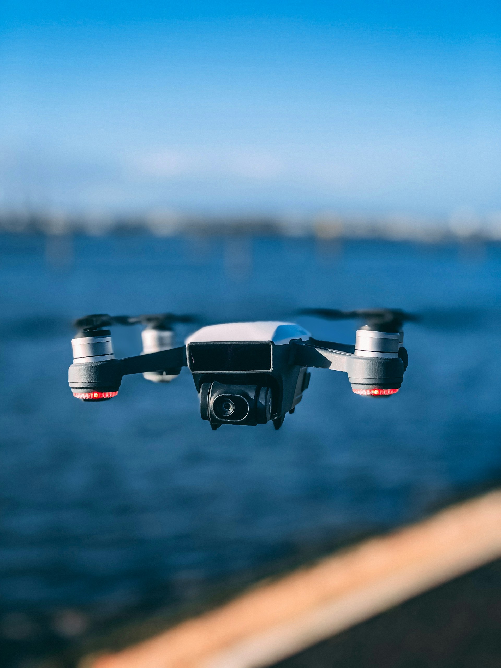 A small white consumer drone with red accents is flying and hovering over a body of water with land in the blurred background.
