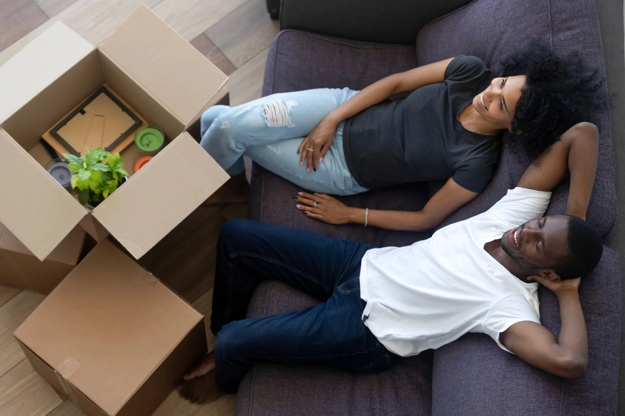 Smiling black couple wearing casual comfortable clothes, sitting on a gray couch, boxes in front of them looks like unpacking stuffs from a move