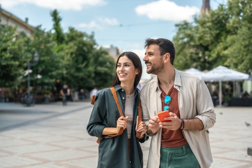 A smiling couple stands outdoors, holding drinks, with trees and a market backdrop under a bright sky.