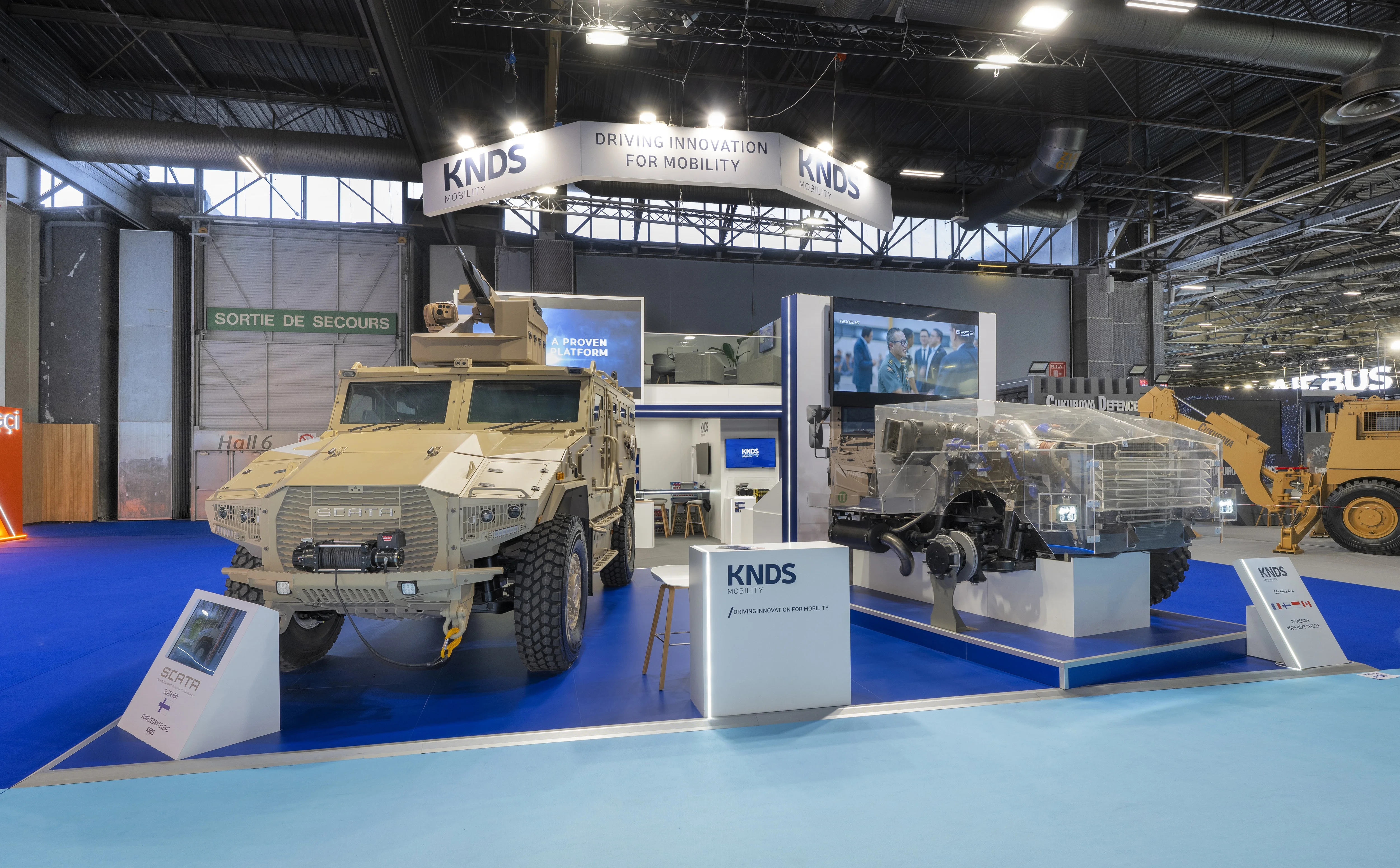 Stand KNDS Mobility conçu et réalisé par Expace à Eurosatory 2026 – espace sur-mesure défense avec blindé SCATA, châssis Celeris écorché, bandeau suspendu « Driving Innovation for Mobility » et lounge à l'étage.