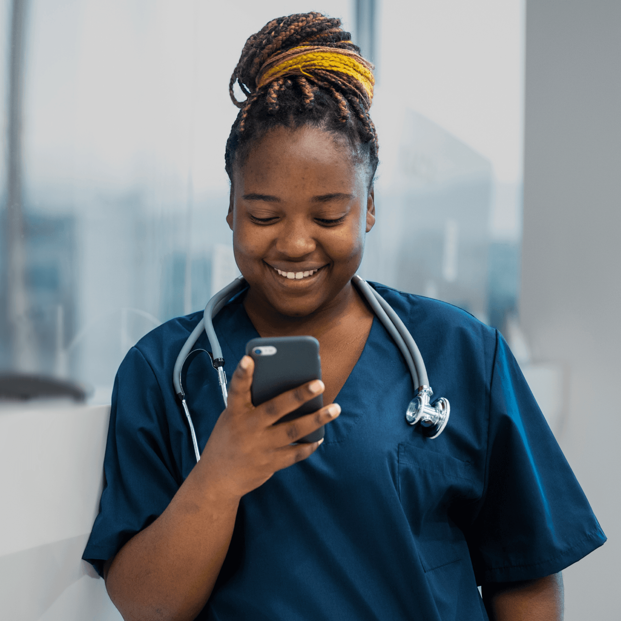 Black female health worker in blue scrubs looking at a phone and smiling, with a background showing a hospital setting.