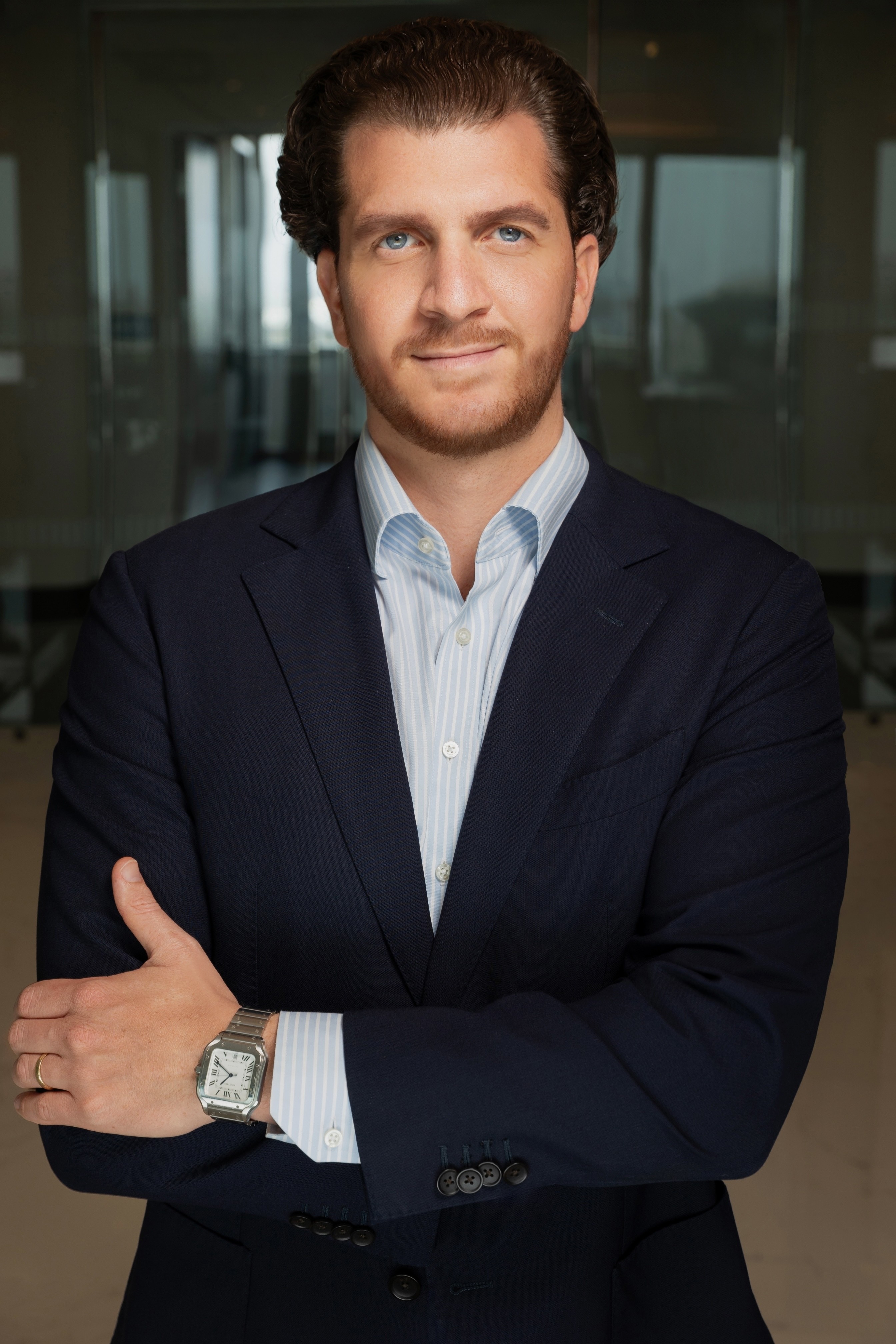 A woman in a black blouse and grey trousers stands confidently with arms crossed, in front of wooden shelves with decorative pottery in a modern office.
