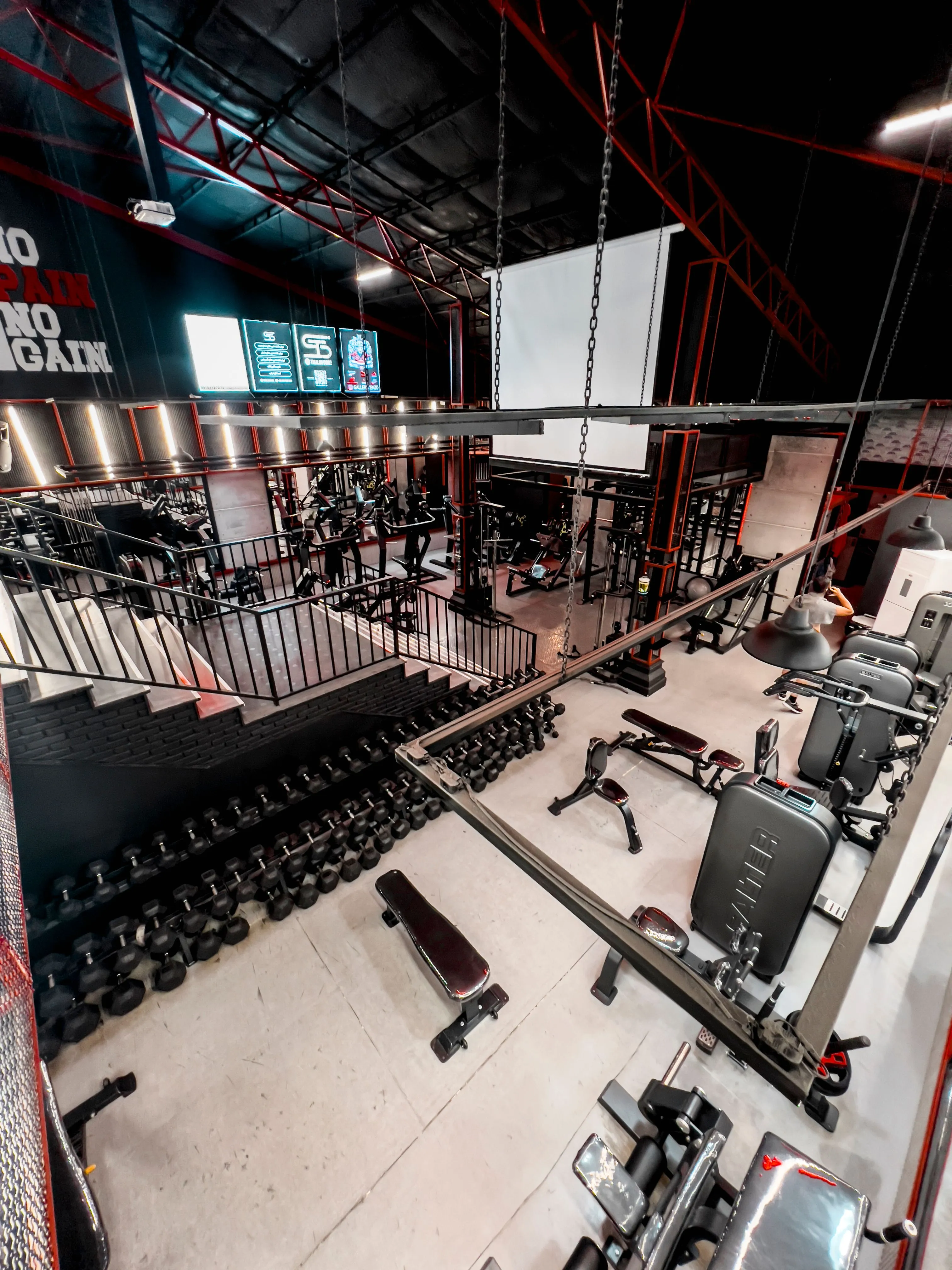 A spacious gym interior featuring multiple rows of black dumbbells, modern weightlifting equipment, and red-accented metal railings, under a high ceiling with industrial lighting.