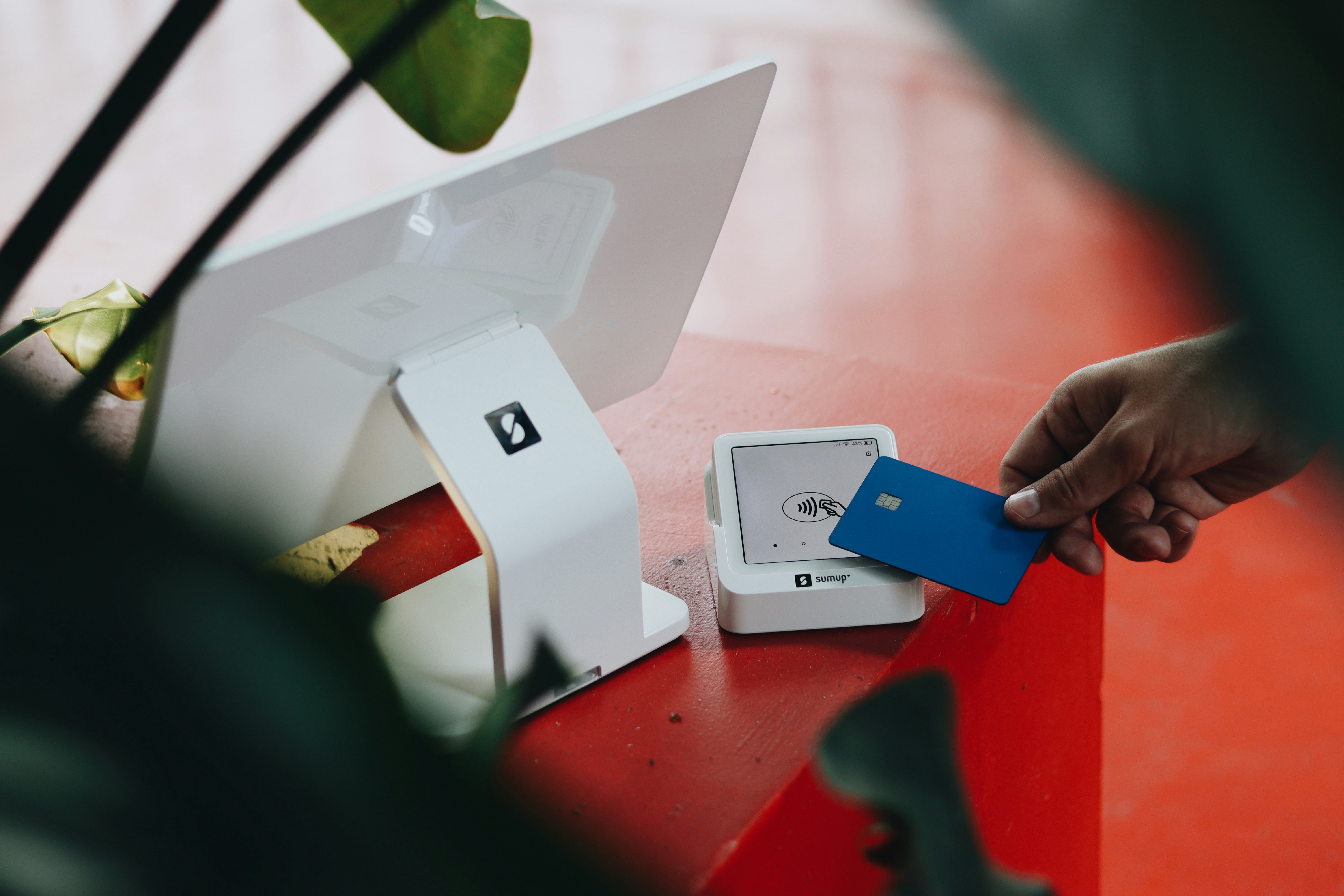 A hand holding a blue payment card near a computer and a white device on a bright orange table.