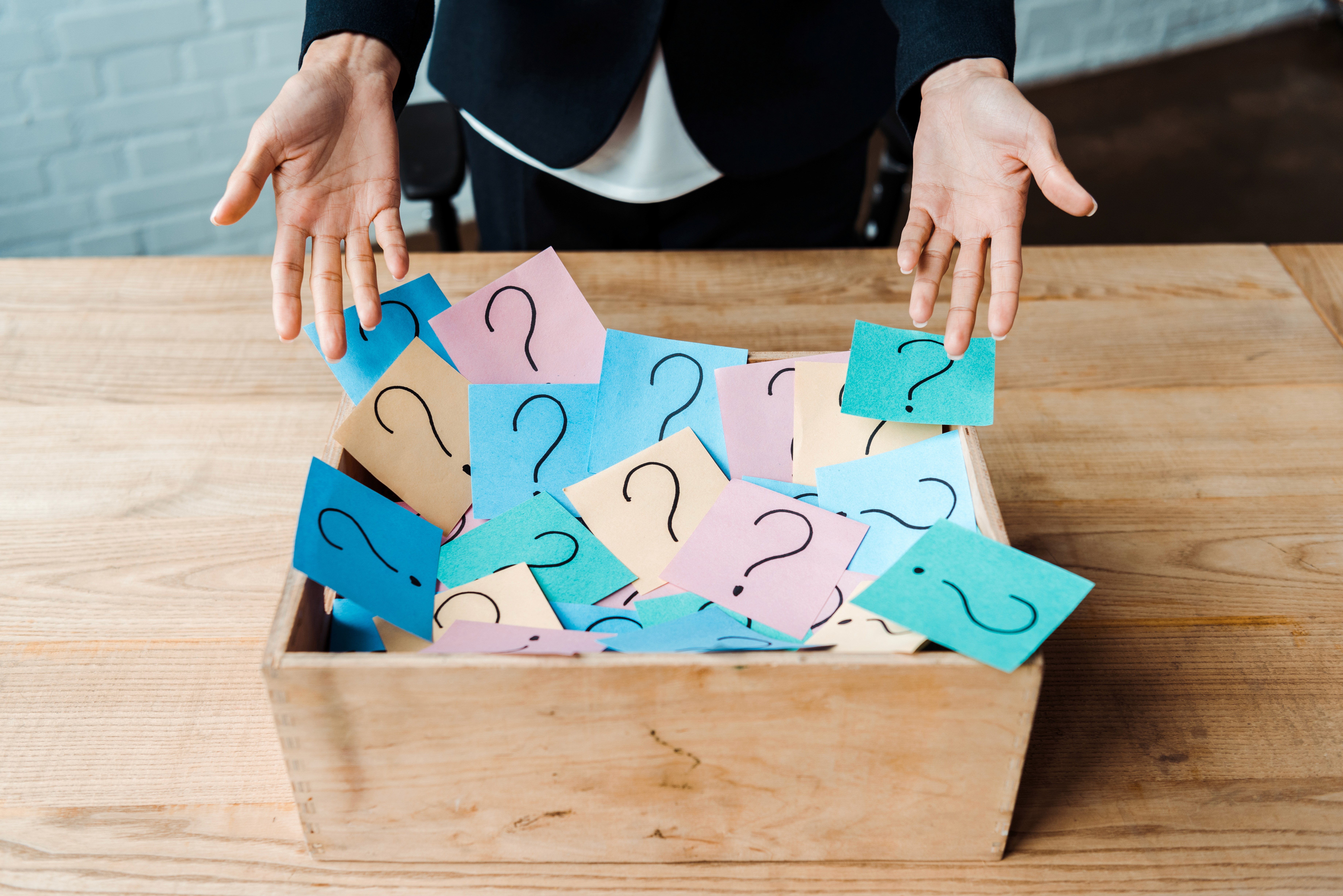 Person gesturing over a wooden box overflowing with colorful sticky notes covered in question marks, representing the confusion and unanswered questions service business owners face when trying to understand marketing concepts like AEO