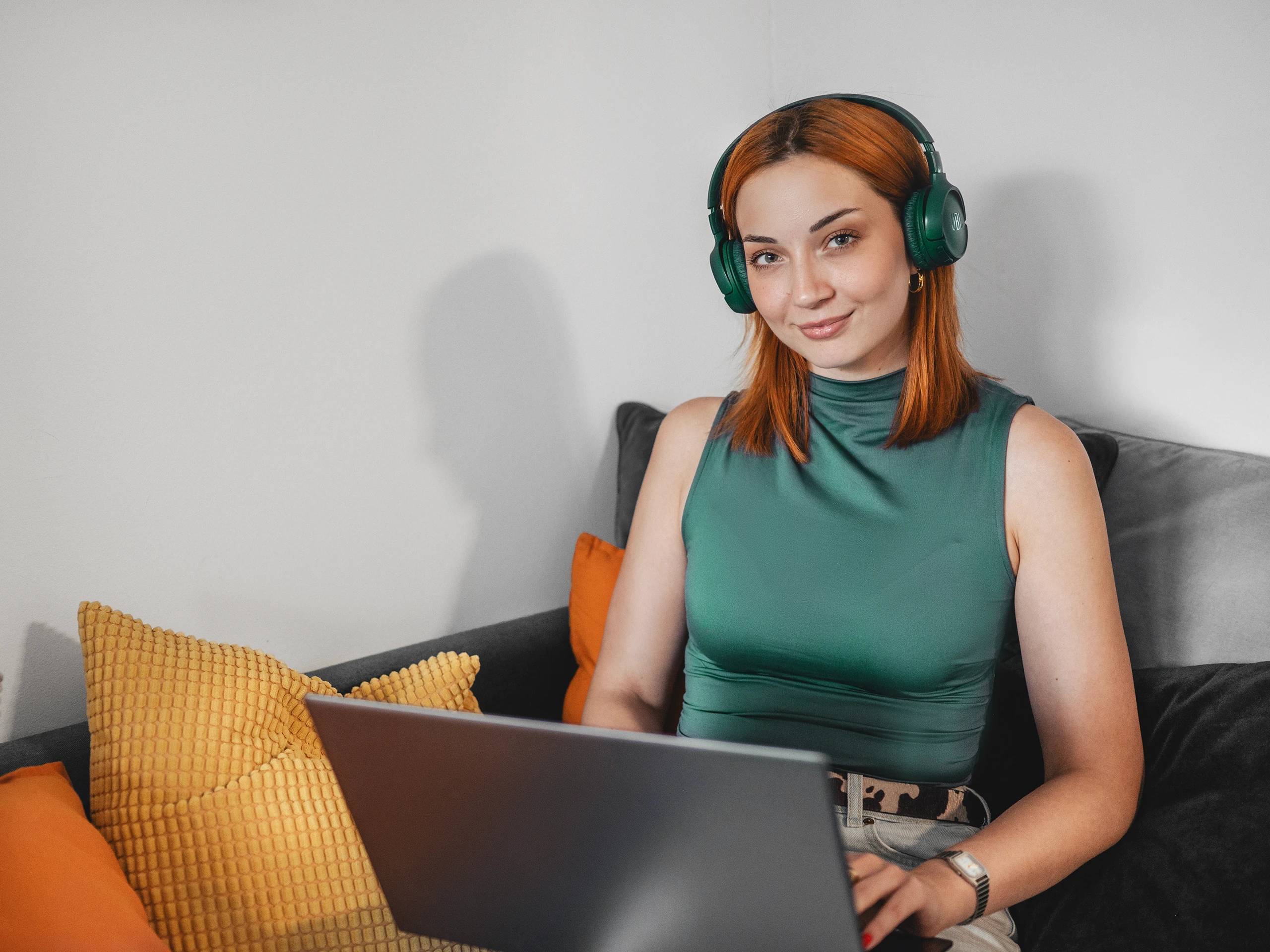 Young woman with red hair wearing green headphones and working on a laptop while sitting on a couch.