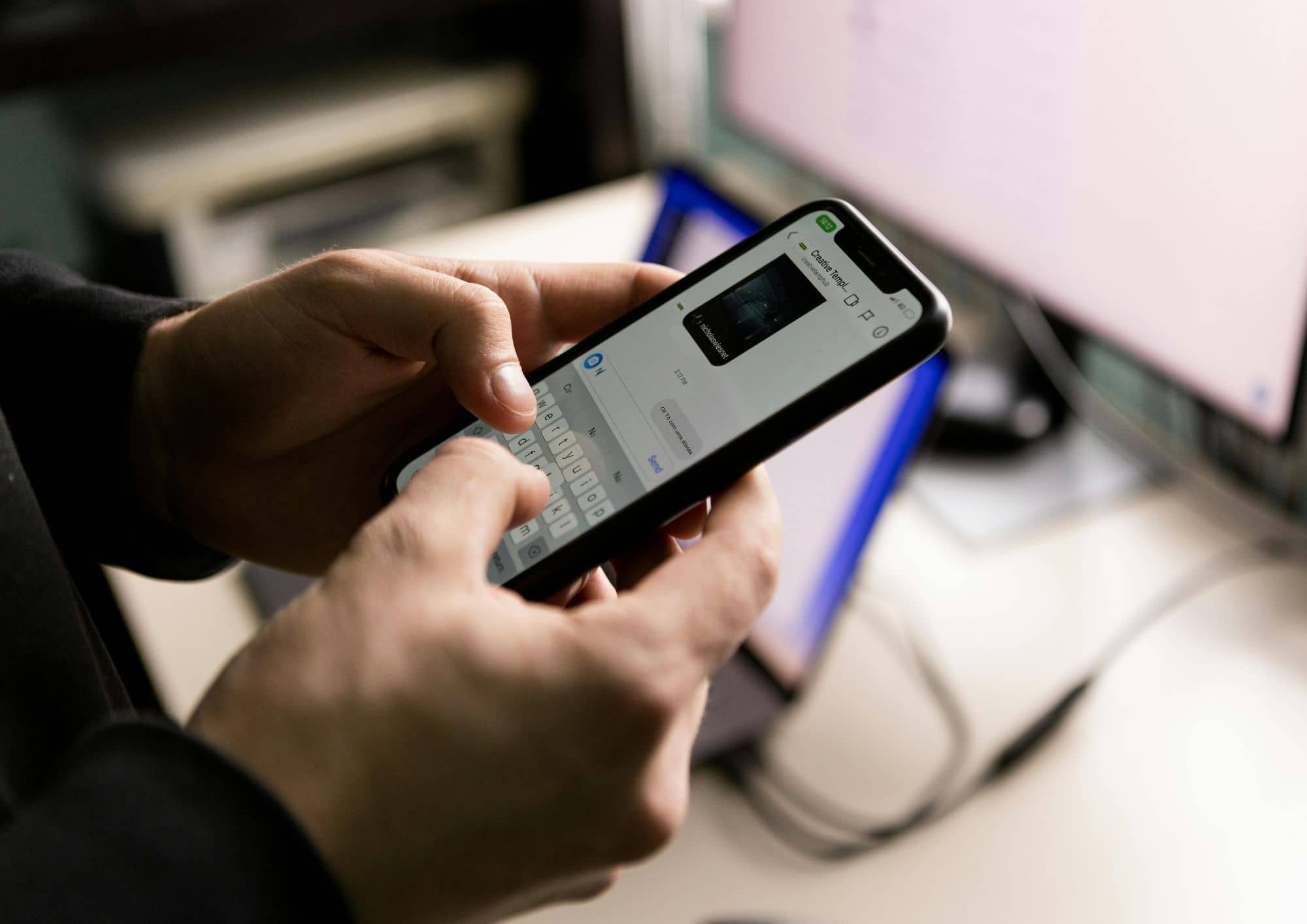 Pessoa segura um smartphone e digita uma mensagem em um aplicativo de conversa. Ao fundo, desfocados, aparecem uma tela de computador e equipamentos de trabalho.