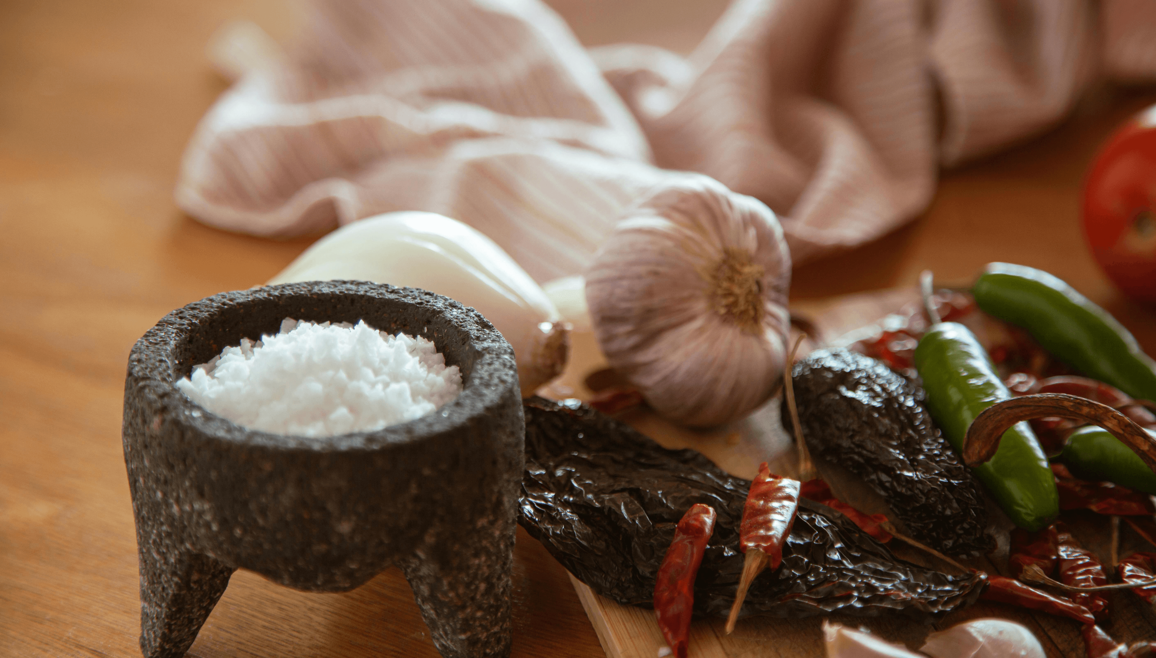 Close-up of a rustic mortar filled with coarse salt, surrounded by fresh garlic, onions, green and red chili peppers, and dried chili peppers on a wooden surface with a striped kitchen cloth in the background.