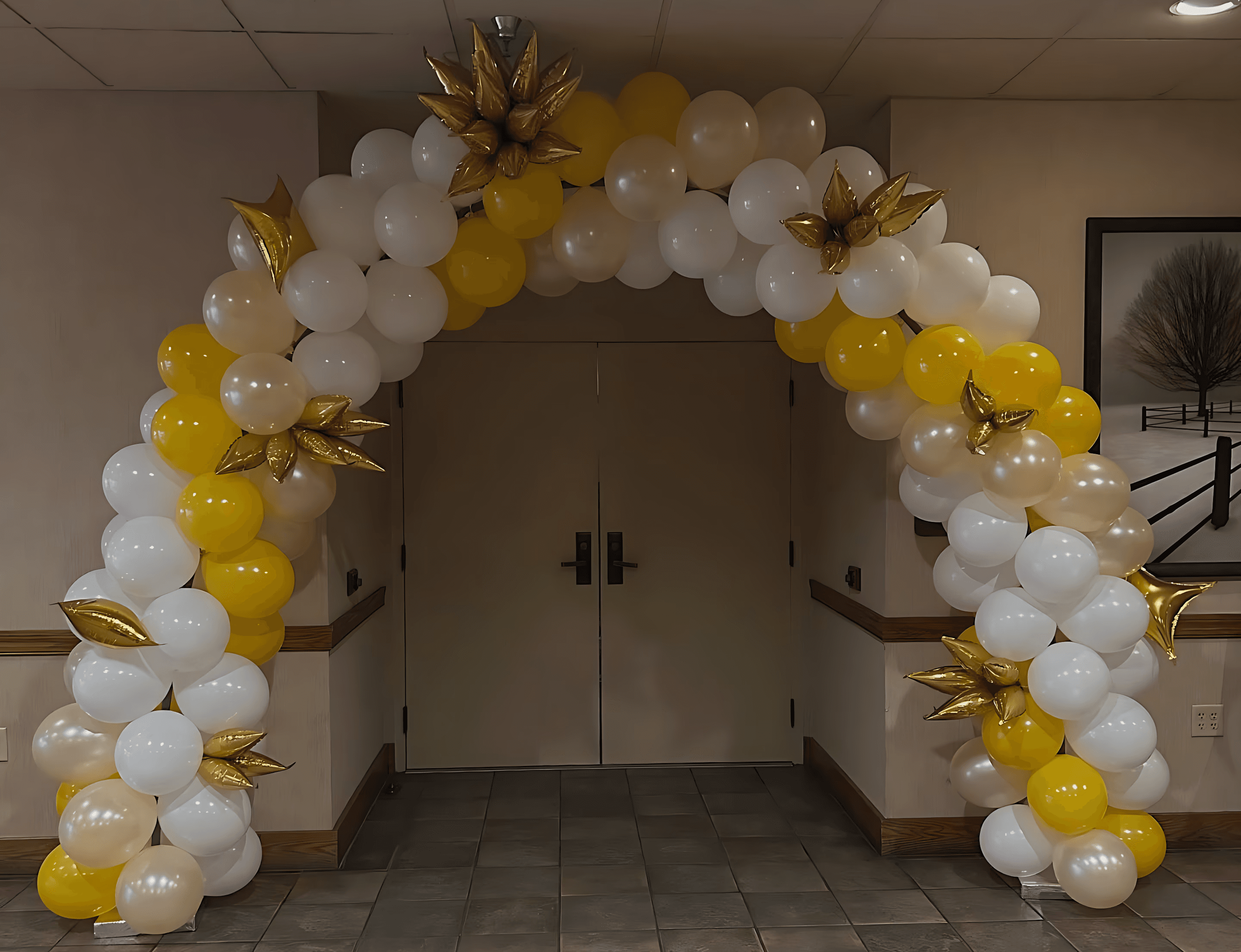 A festive balloon arch made of white, yellow, and gold balloons, adorned with gold star decorations, frames a doorway in a tiled hallway.