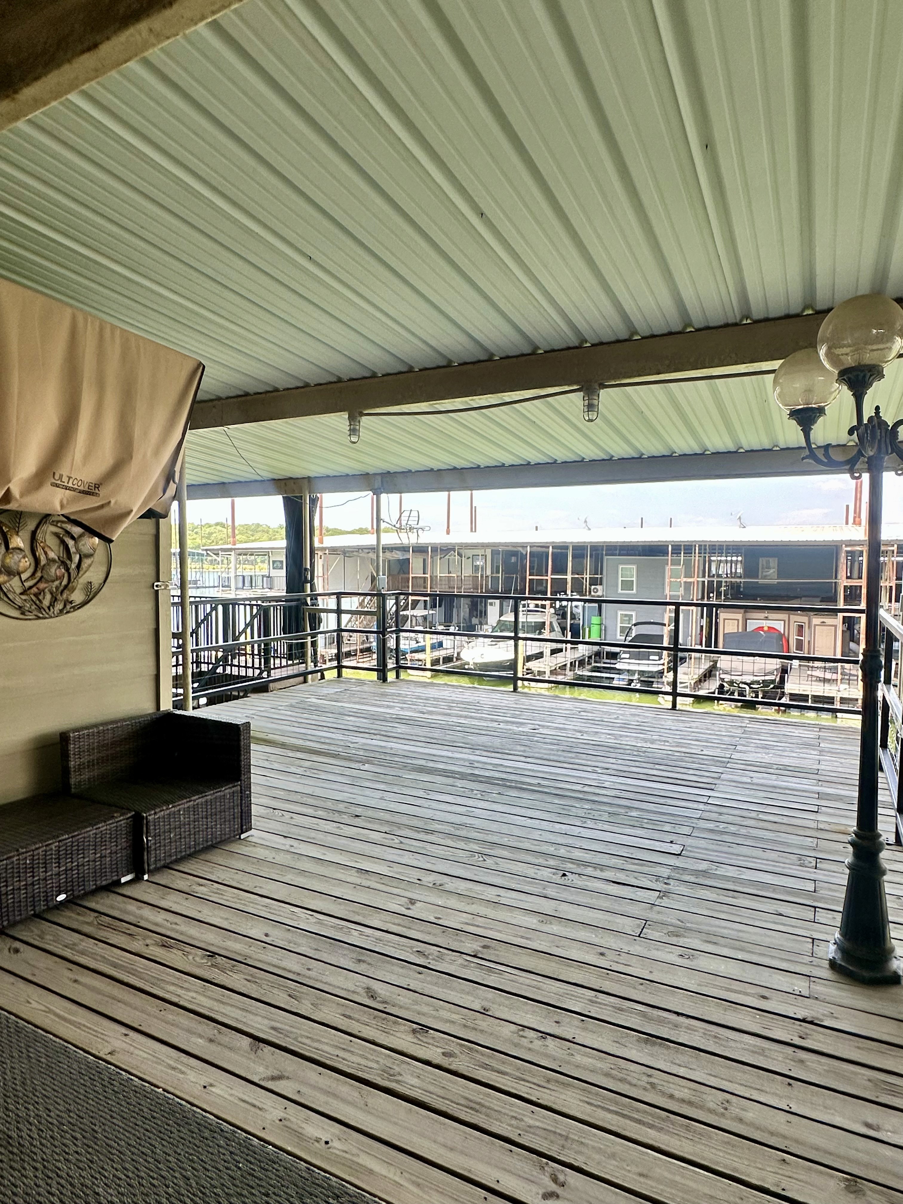 Spacious covered wooden deck overlooking a marina, with wicker furniture on the left and a vintage streetlamp on the right.