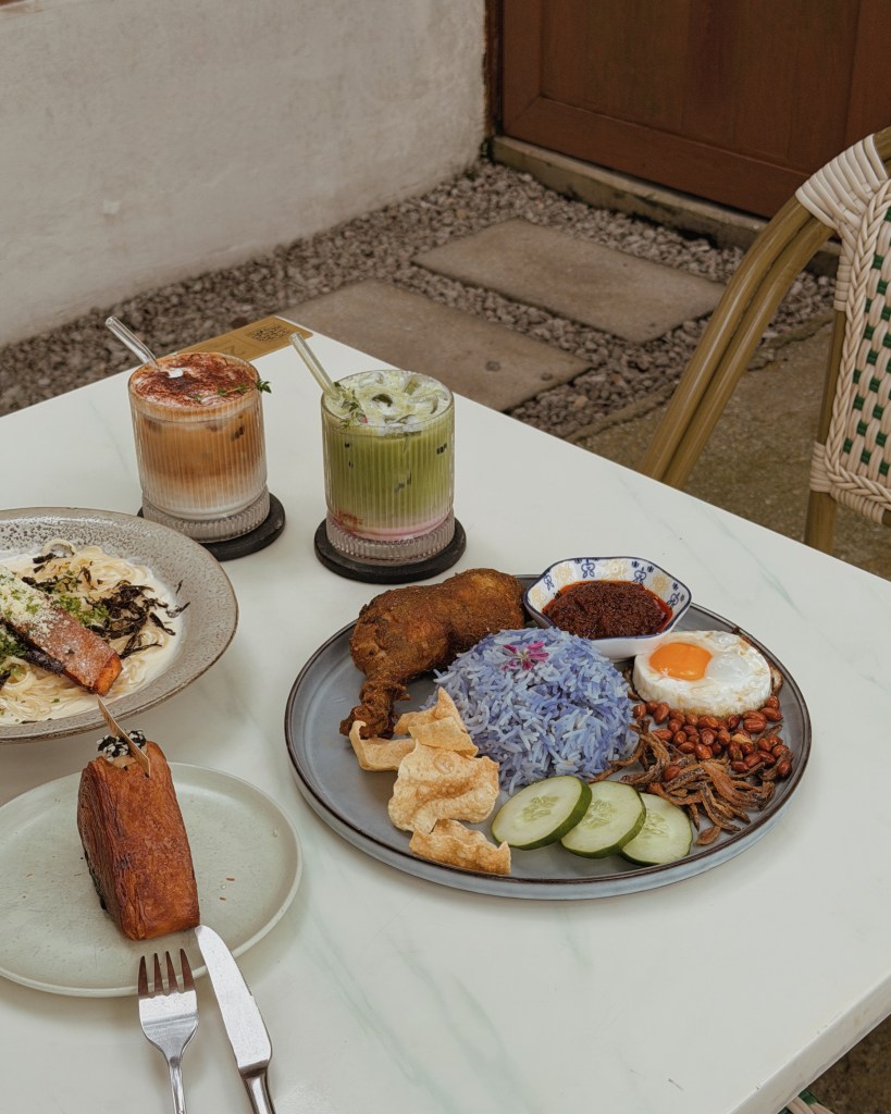 A food spread featuring a plate of Nasi Lemak with blue rice, fried chicken, peanuts, cucumber slices, fried egg, and sambal, accompanied by a side of crackers. In the background, two glasses of iced drinks and a plate of pasta with salmon are visible.