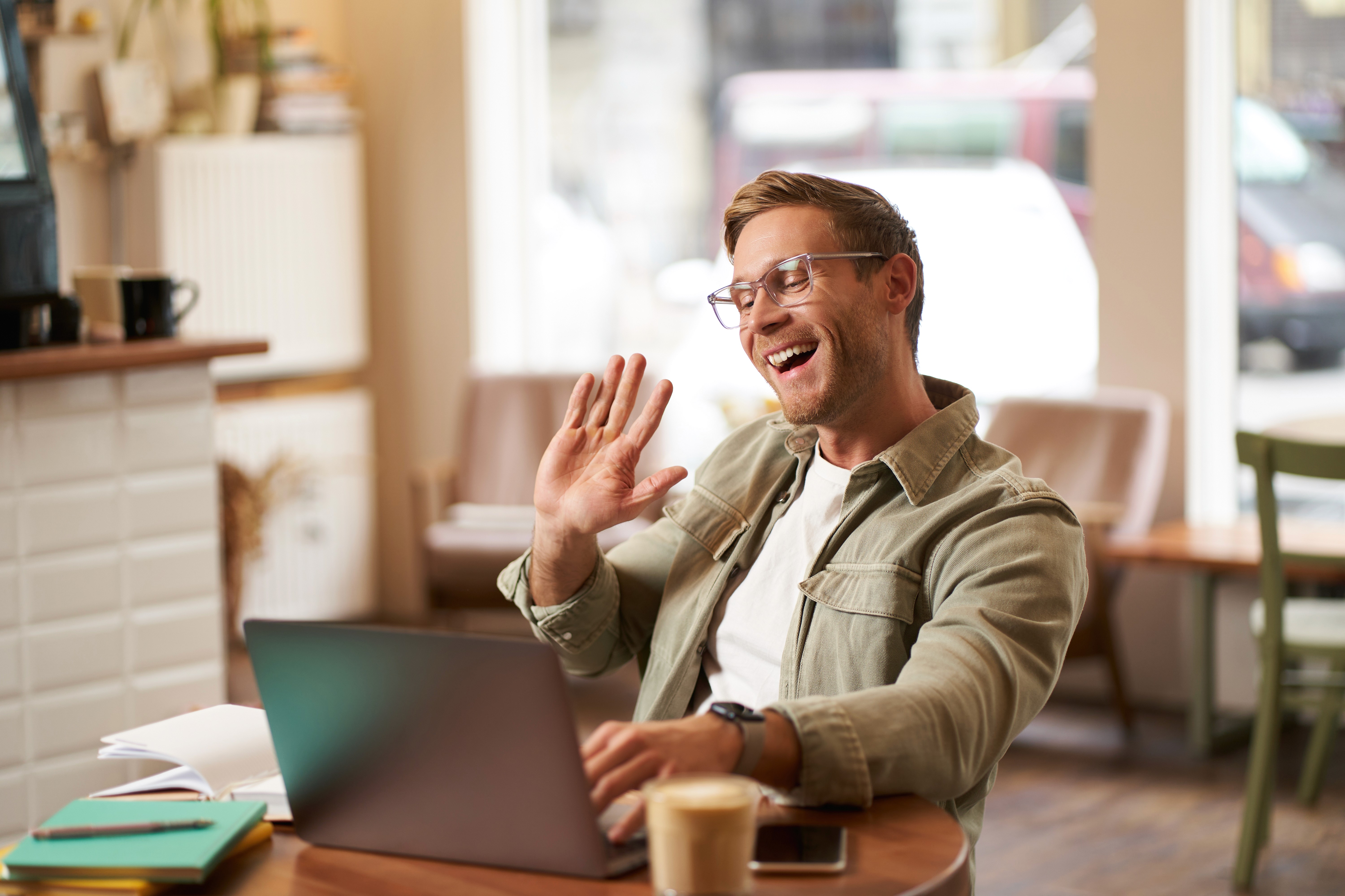 Junger Mann mit Brille sitzt an einem Tisch im Café vor einem Laptop und winkt mit der rechten Hand