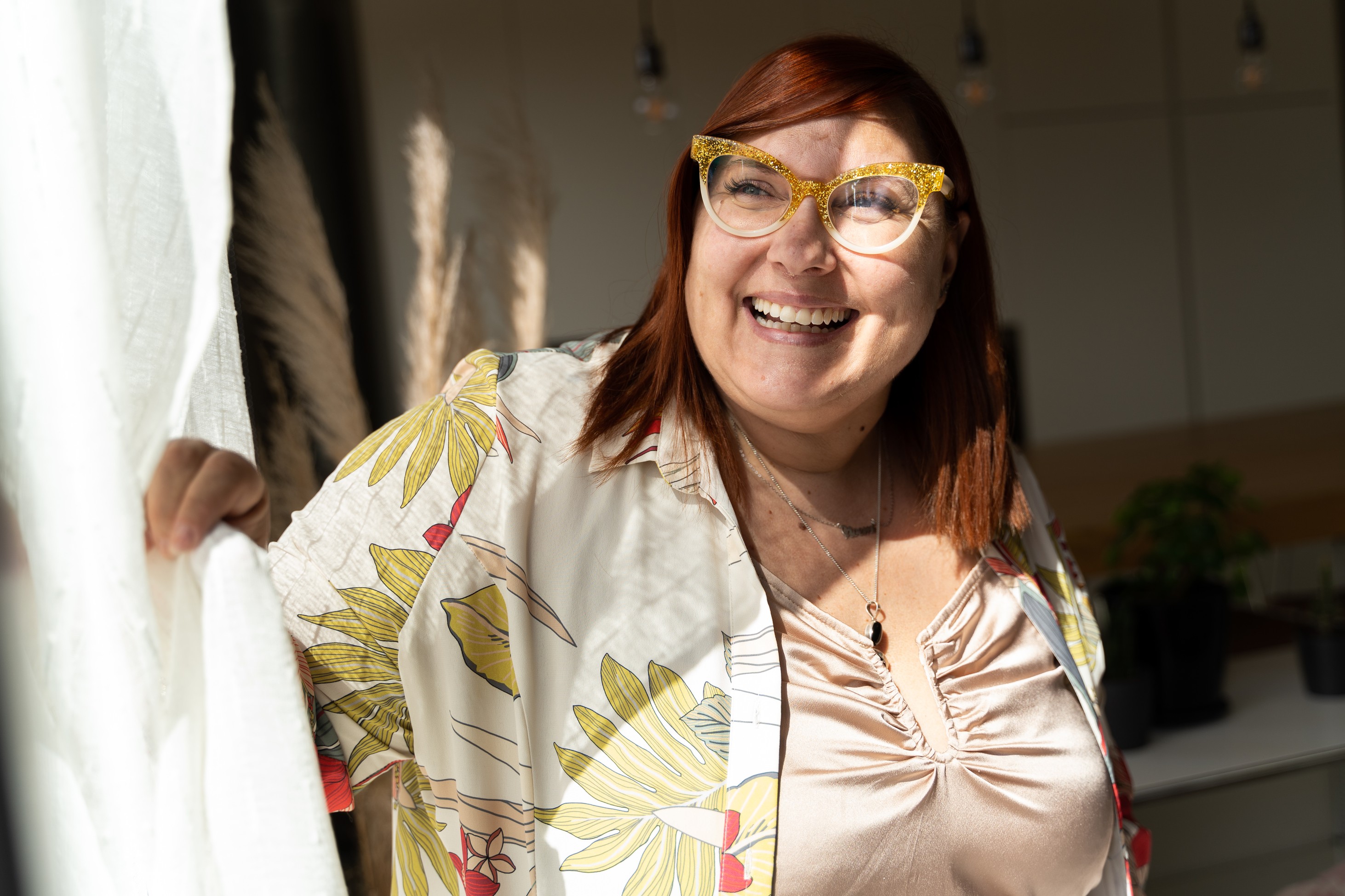 A retiree smiles by a sunlit window, holding a sheer curtain, wearing yellow glasses and a floral jacket