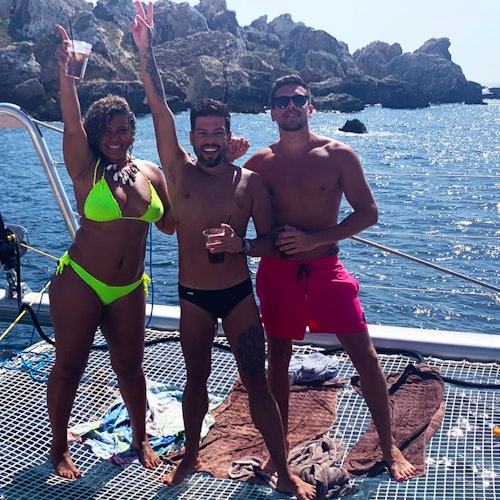 Three people in swimwear stand on a boat deck by the sea, smiling and holding drinks with rocky cliffs in the background.