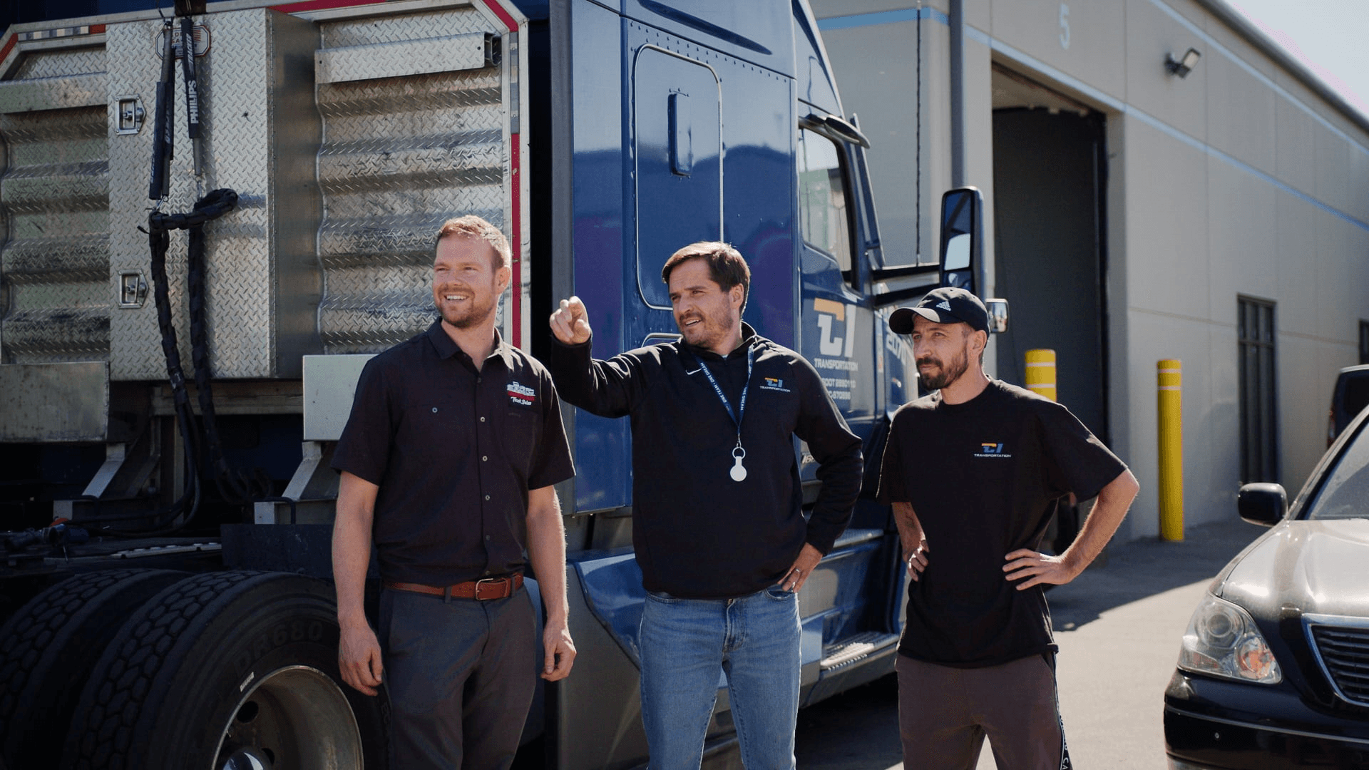 Three transportation professionals stand beside a blue semi-truck outside a logistics facility.