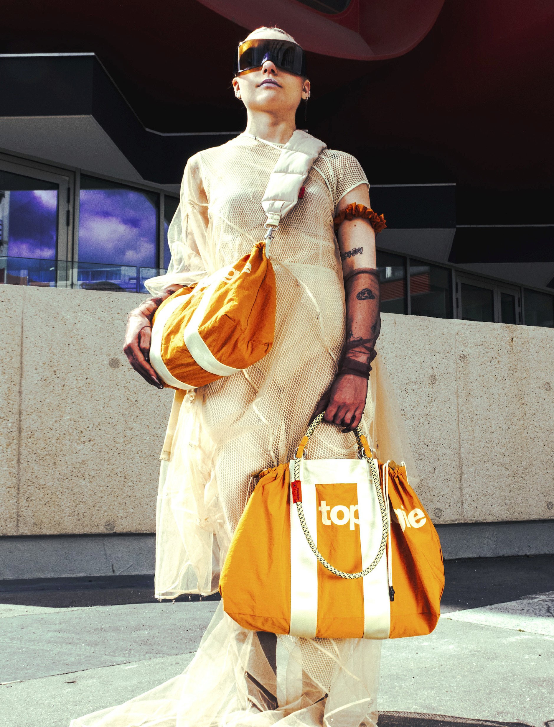 Une femme avec des lunettes retr-futuriste et une robe blanche porte deux sacs orange de la marque topologie