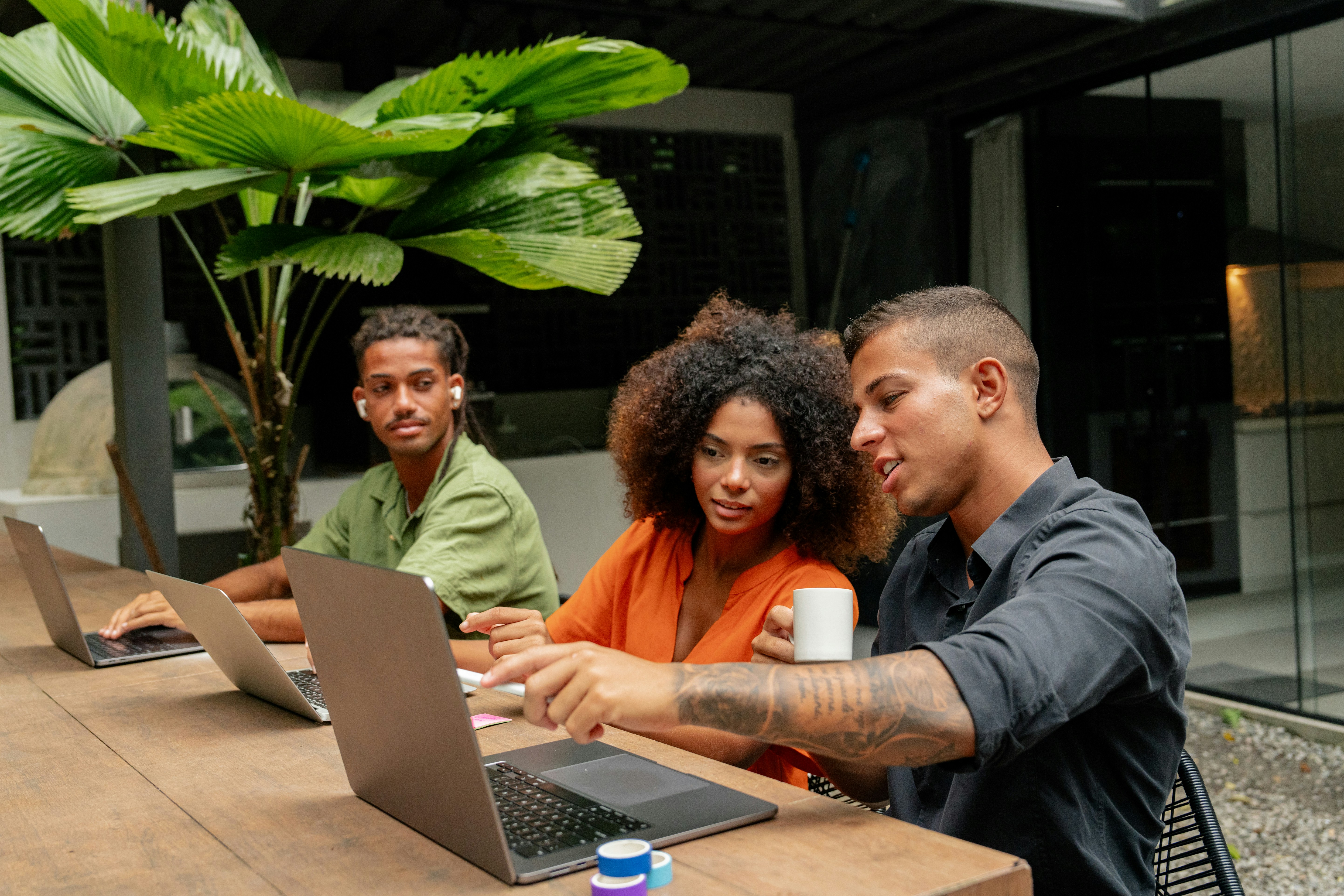 Three people collaborate around a laptop at a table, surrounded by green plants in a modern workspace.