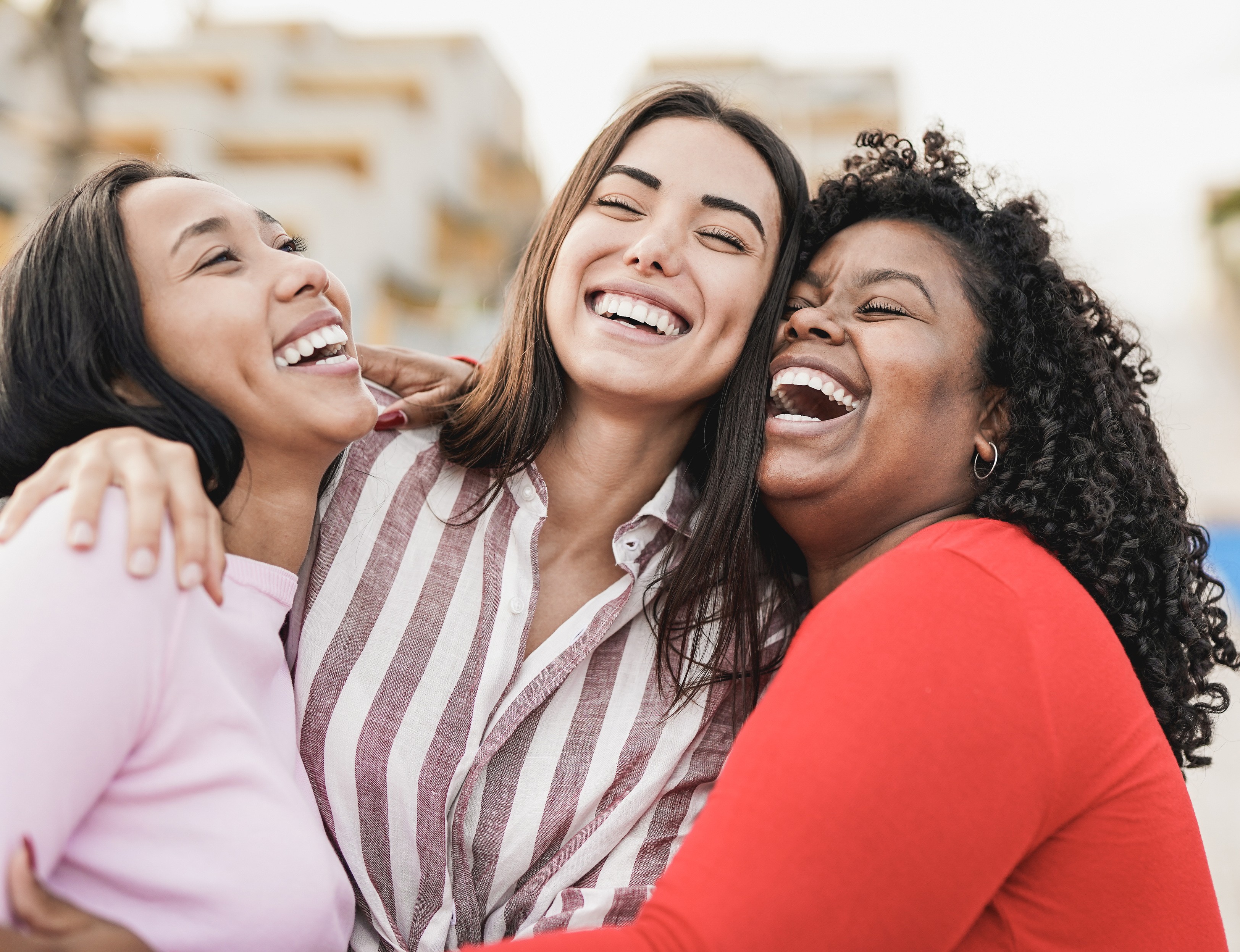 A imagem mostra três mulheres abraçadas, sorrindo e rindo juntas. Elas parecem estar ao ar livre, com uma paisagem urbana desfocada ao fundo. A mulher à esquerda veste uma blusa rosa clara, enquanto a mulher ao centro usa uma camisa listrada em vermelho e branco. A mulher à direita está usando uma blusa vermelha e tem cabelos cacheados. A atmosfera da foto transmite alegria, amizade e companheirismo, com as três mulheres compartilhando um momento descontraído e divertido.