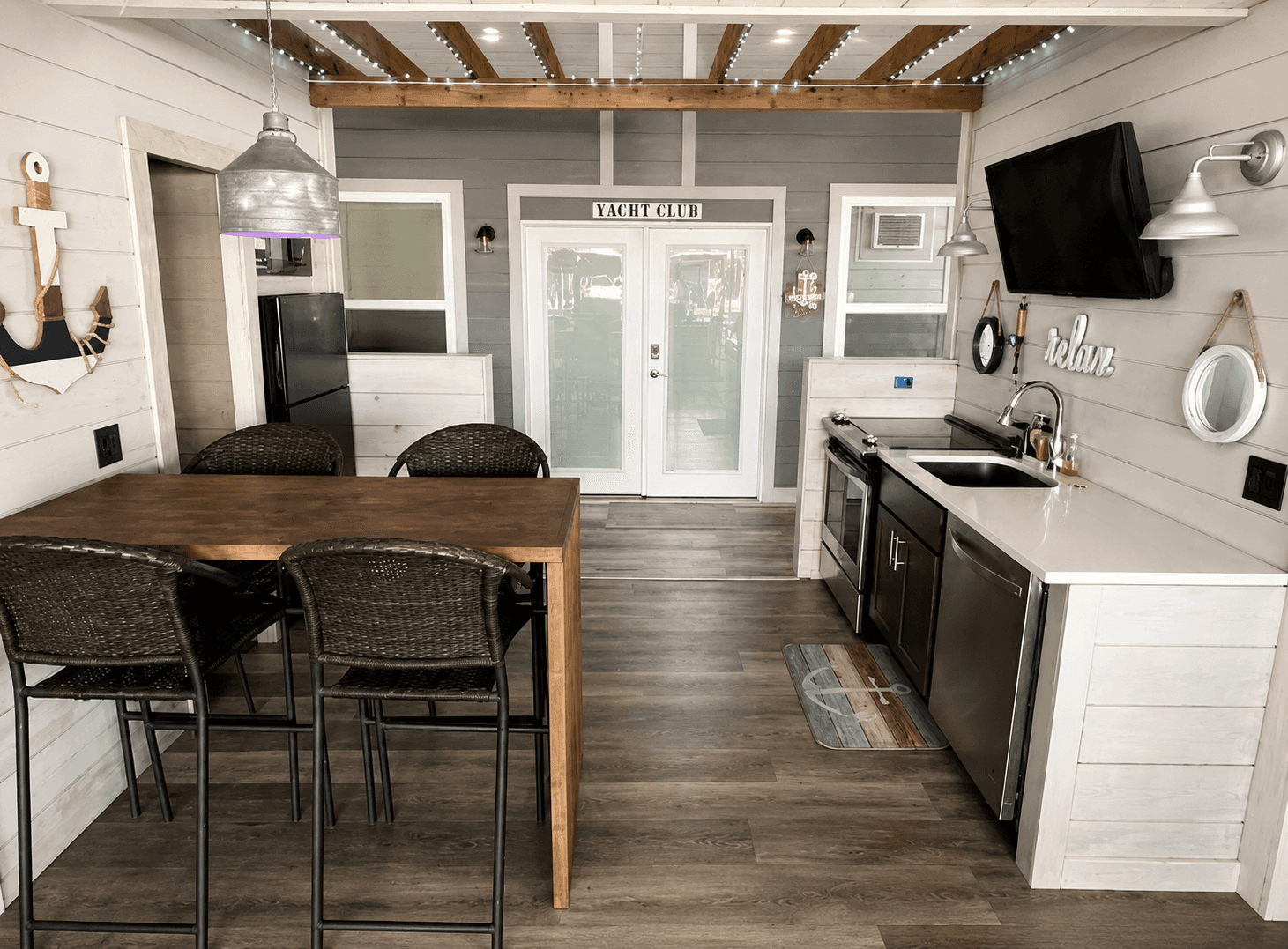Modern coastal-themed kitchen with wooden beams, a wooden dining table with wicker chairs, stainless steel appliances, a farmhouse sink, and a nautical décor element next to glass doors labeled "YACHT CLUB."