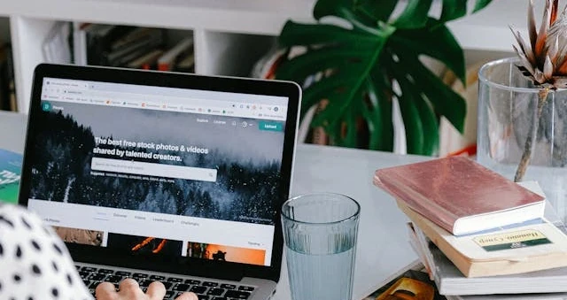 Mesa de trabalho com laptop aberto, pilha de livros e plantas, com uma pessoa digitando