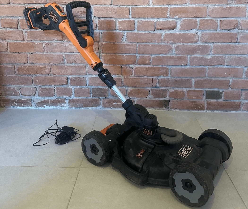 Tondeuse et coupe-bordure sans fil 2-en-1 BLACK+DECKER orange et noire avec chargeur, posée sur un sol en céramique devant un mur de briques à Montréal.