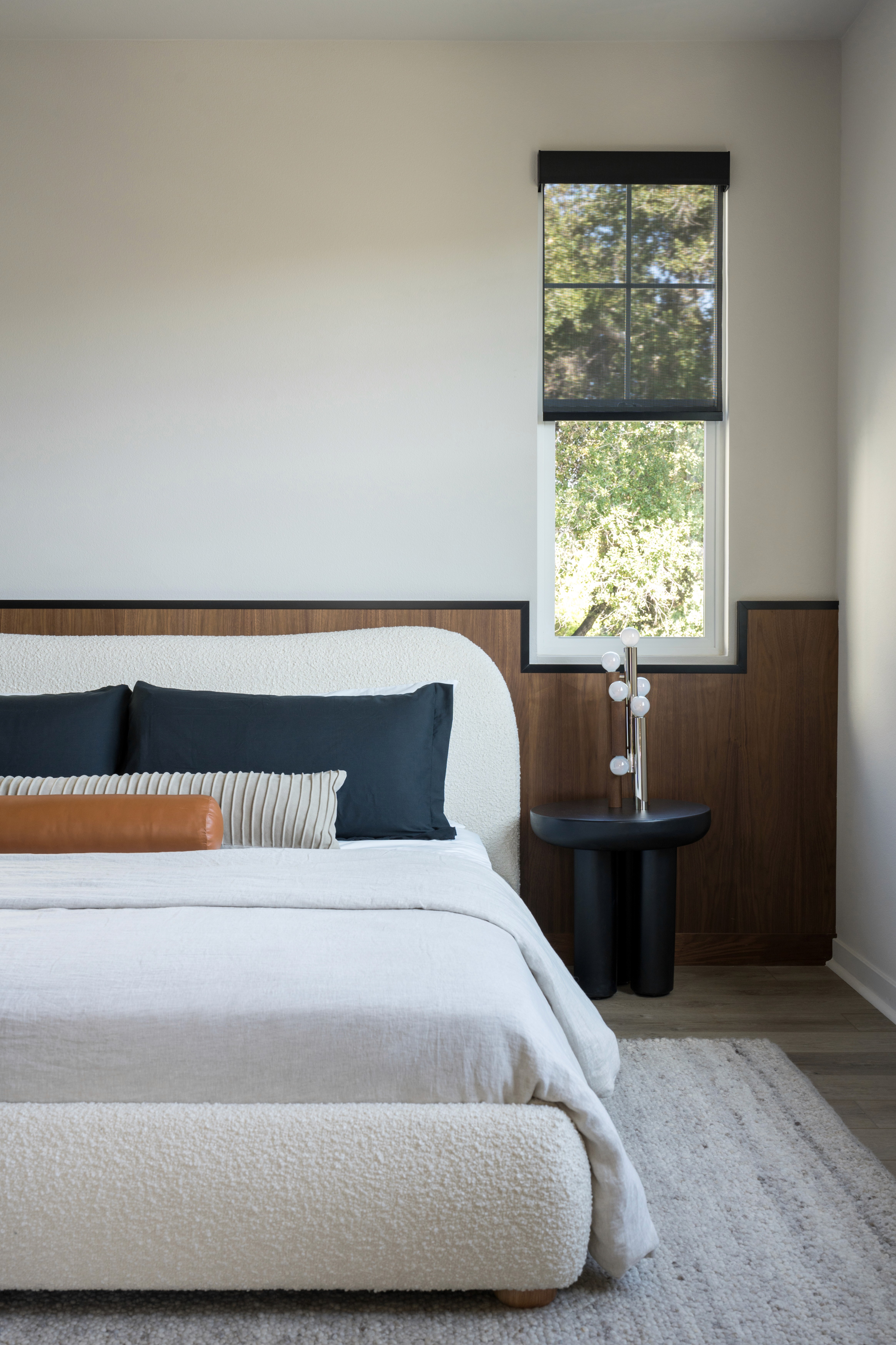 Moody bedroom with boucle bed frame, dark blue, leather and grey accent pillows, grey and white rug, black side table, and black lined, warm wood wainscoting by Jubilee Interiors in Arcadia, California