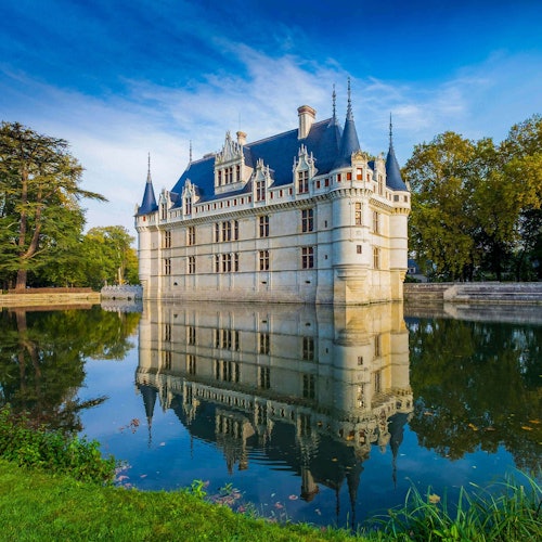 Un grand château avec plusieurs tours pointues se reflète dans un fossé paisible environnant, avec des arbres en arrière-plan sous un ciel bleu.