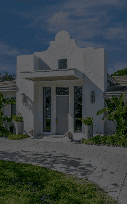 Modern Southwest-style home with clean white stucco exterior and distinctive parapet roofline. Features dramatic front entryway with tall glass doors flanked by lantern sconces, large garage, and minimalist drought-resistant landscaping with tropical accent plants. Gray concrete paver driveway complements the architectural style against deep blue sky