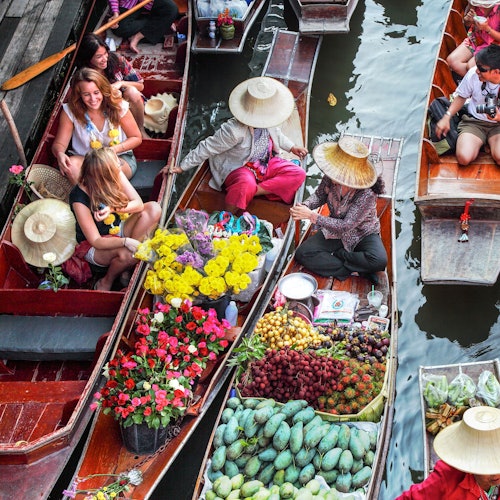 사람들은 형형색색의 꽃들과 다양한 과일로 가득 찬 나무 배에 올라 떠 있는 시장을 거닙니다. 한 배에는 쇼핑하는 여성들이 있습니다.