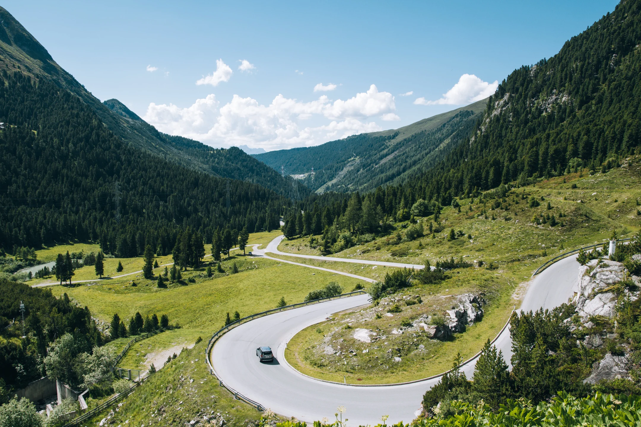 A winding mountain road in Switzerland surrounded by lush green hills and forests under a clear blue sky.