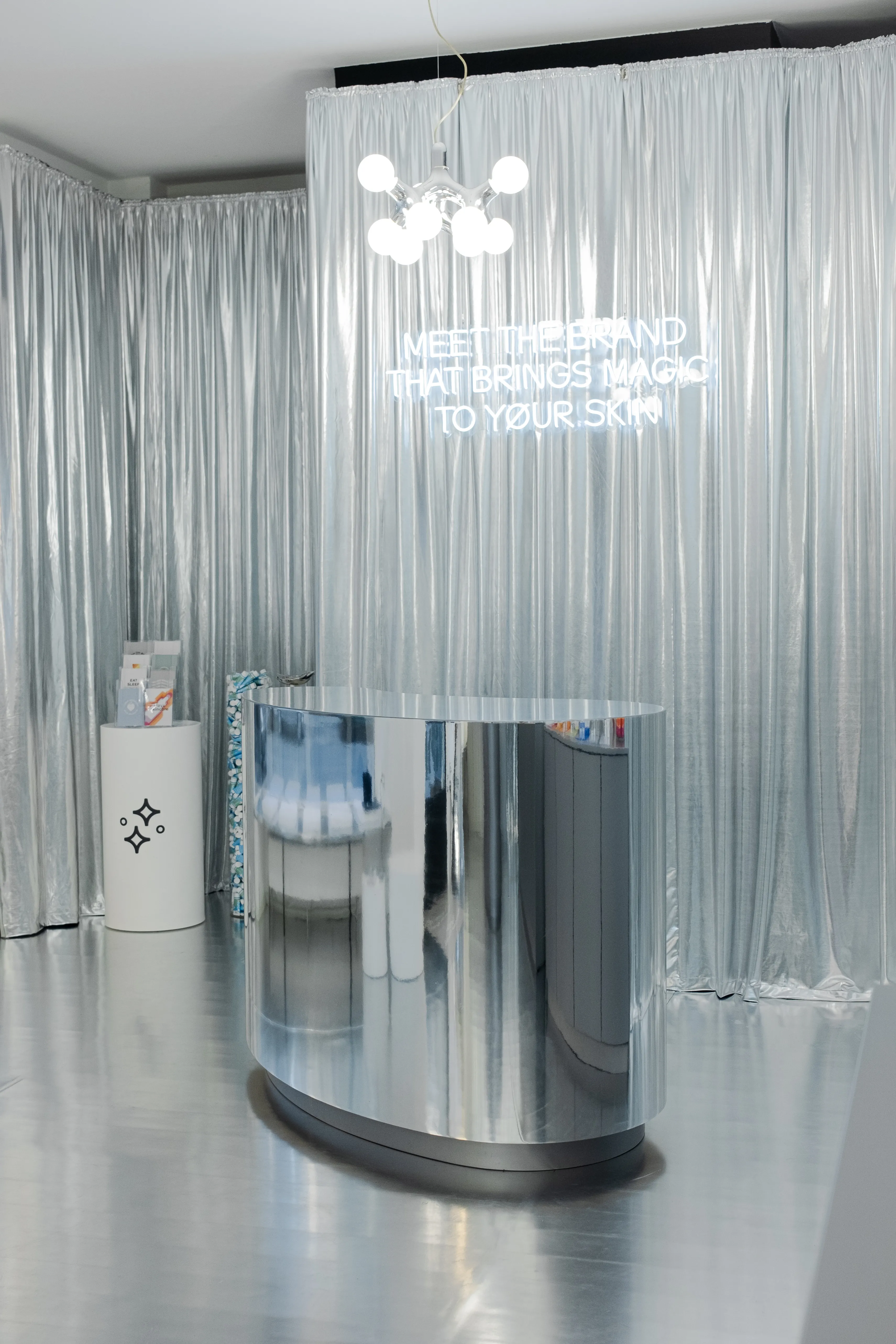 Chrome counter under the neon line Meet the Brand That Brings Magic to Your Skin, framed by silver curtains.