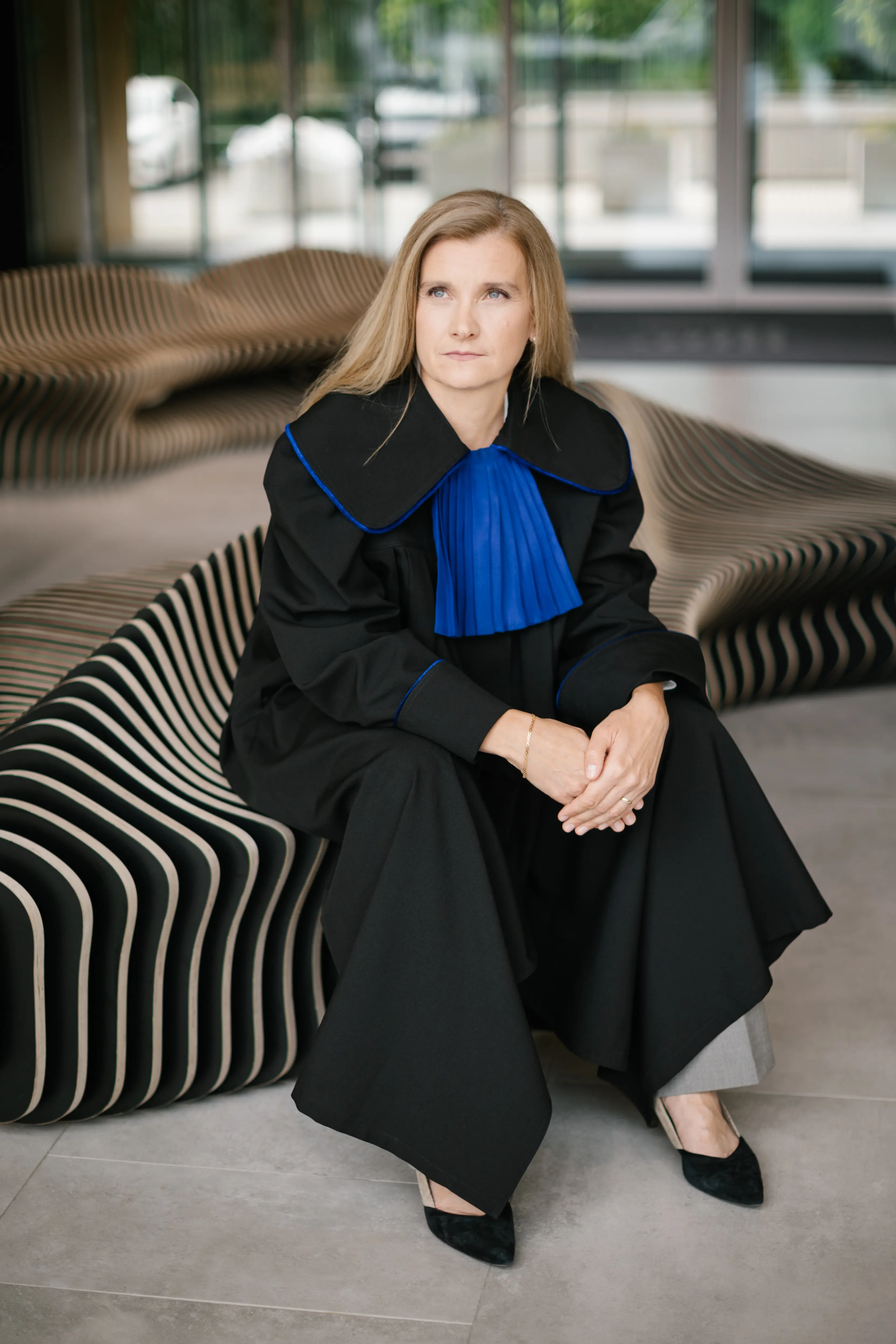 obieta o blond włosach, ubrana w czarną szatę z niebieskim plisowanym kołnierzem, siedzi na nowoczesnej, pasiastą ławce w pomieszczeniu, patrząc lekko w bok z zamyślonym wyrazem twarzy.