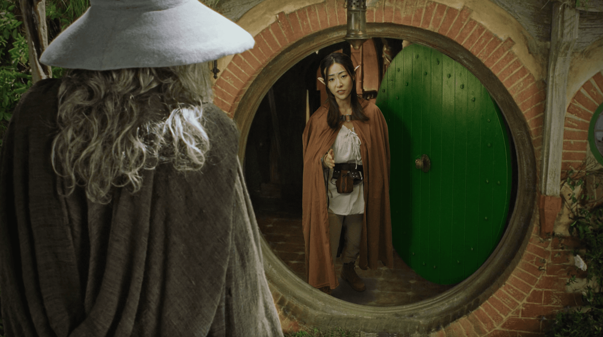 A tall grey-cloaked figure from behind at the round green door of Bag End.